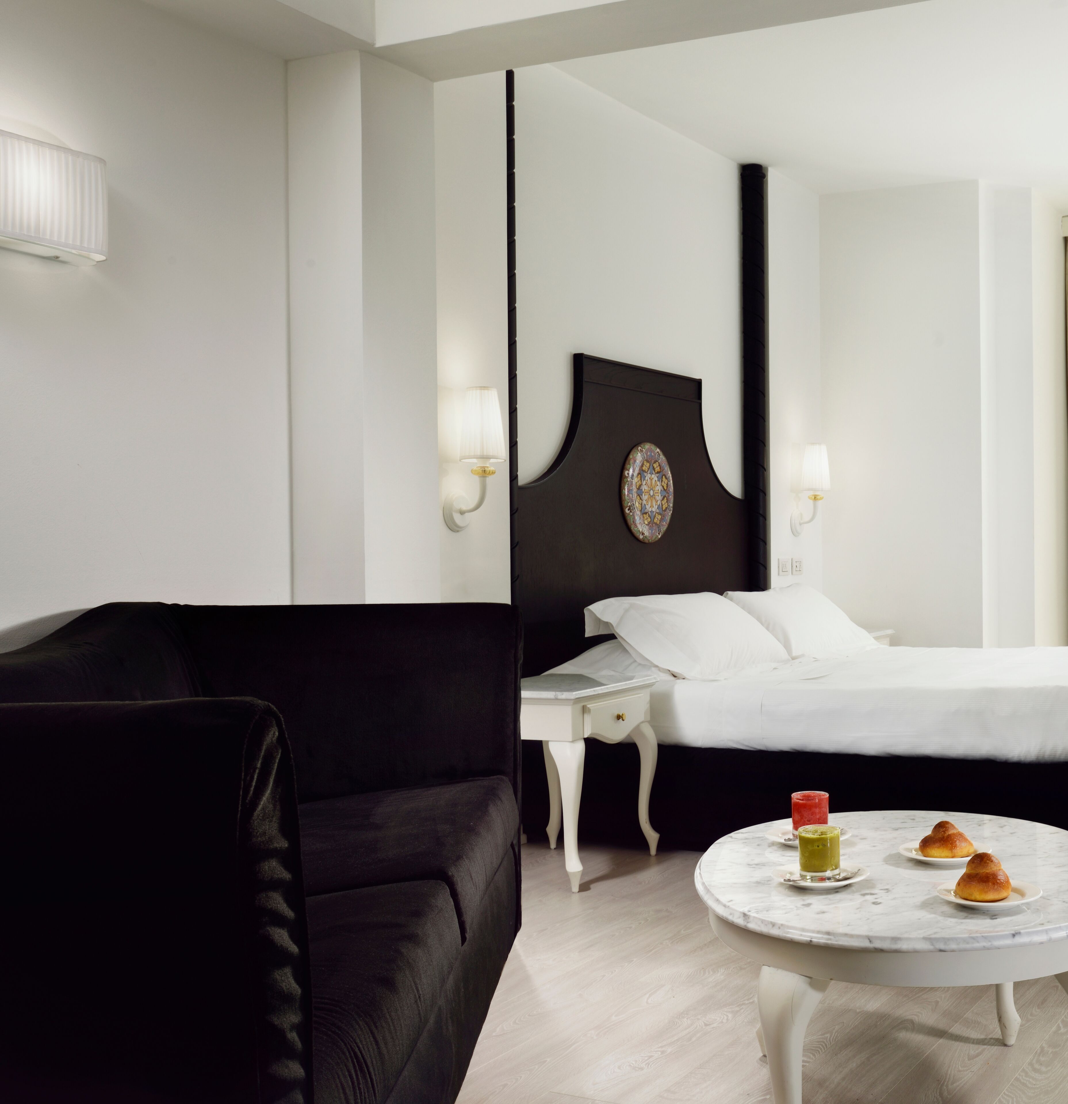 Une chambre d’hôtel avec un canapé noir, un lit blanc avec du linge de lit blanc, une table ronde en marbre sur laquelle se trouvent une boisson et des pâtisseries, et un grand miroir au mur.
