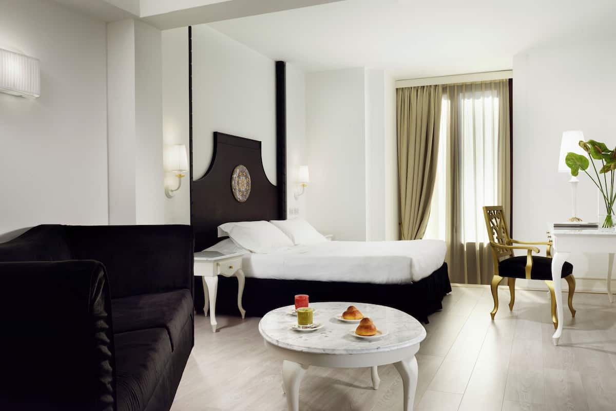 A hotel room with a bed, a black sofa, a round table with pastries, a desk, and a chair.