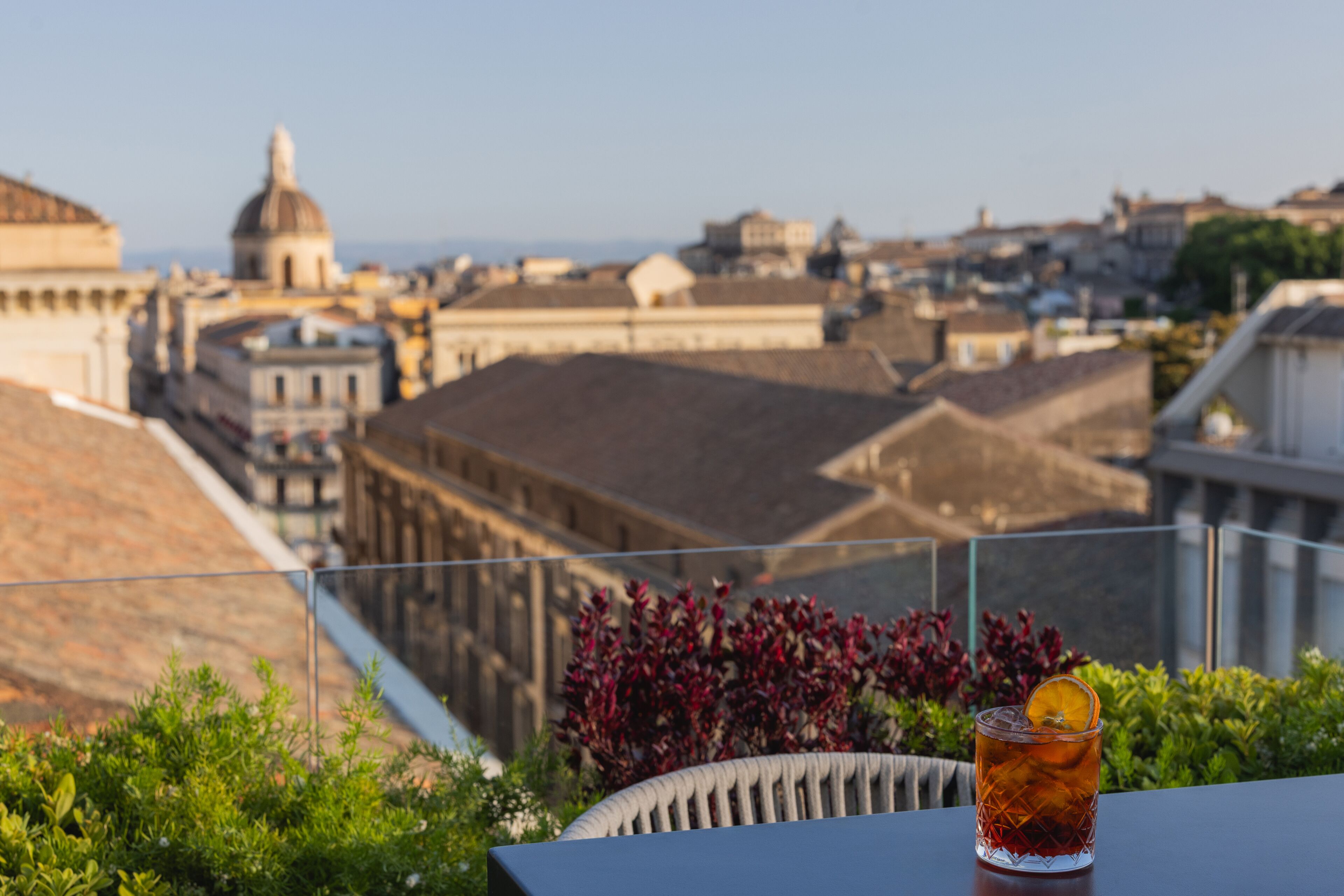 Un balcon avec une boisson, de la verdure et une vue sur la ville en arrière-plan.