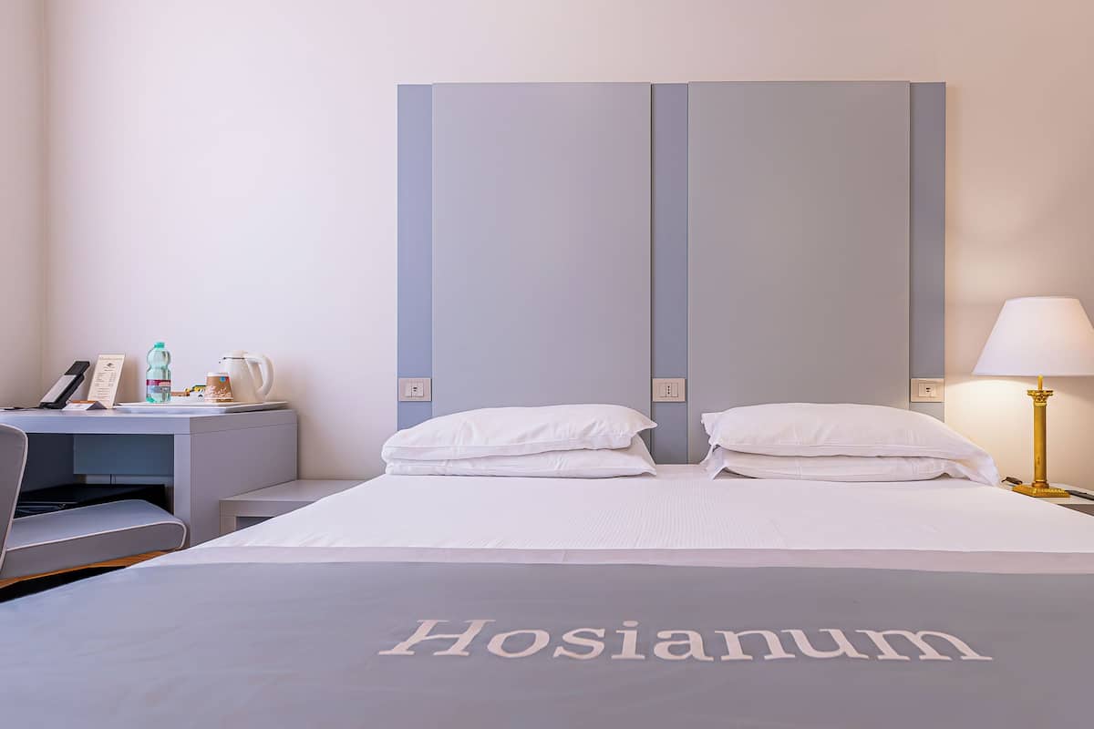 A hotel room with a bed labeled 'Hosianum,' a desk with a chair, a bedside table with a lamp, and a wall-mounted phone.