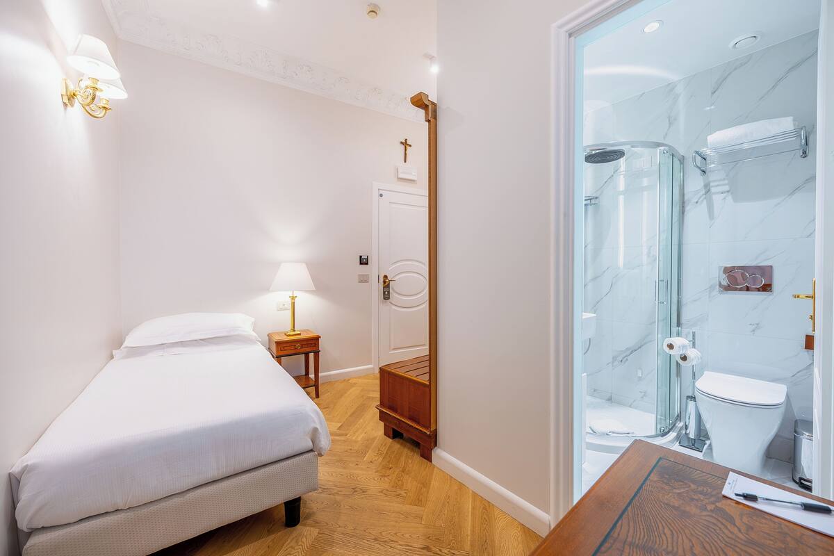 A hotel room with a bed, bedside table, lamp, wooden floor, and a glass shower enclosure.