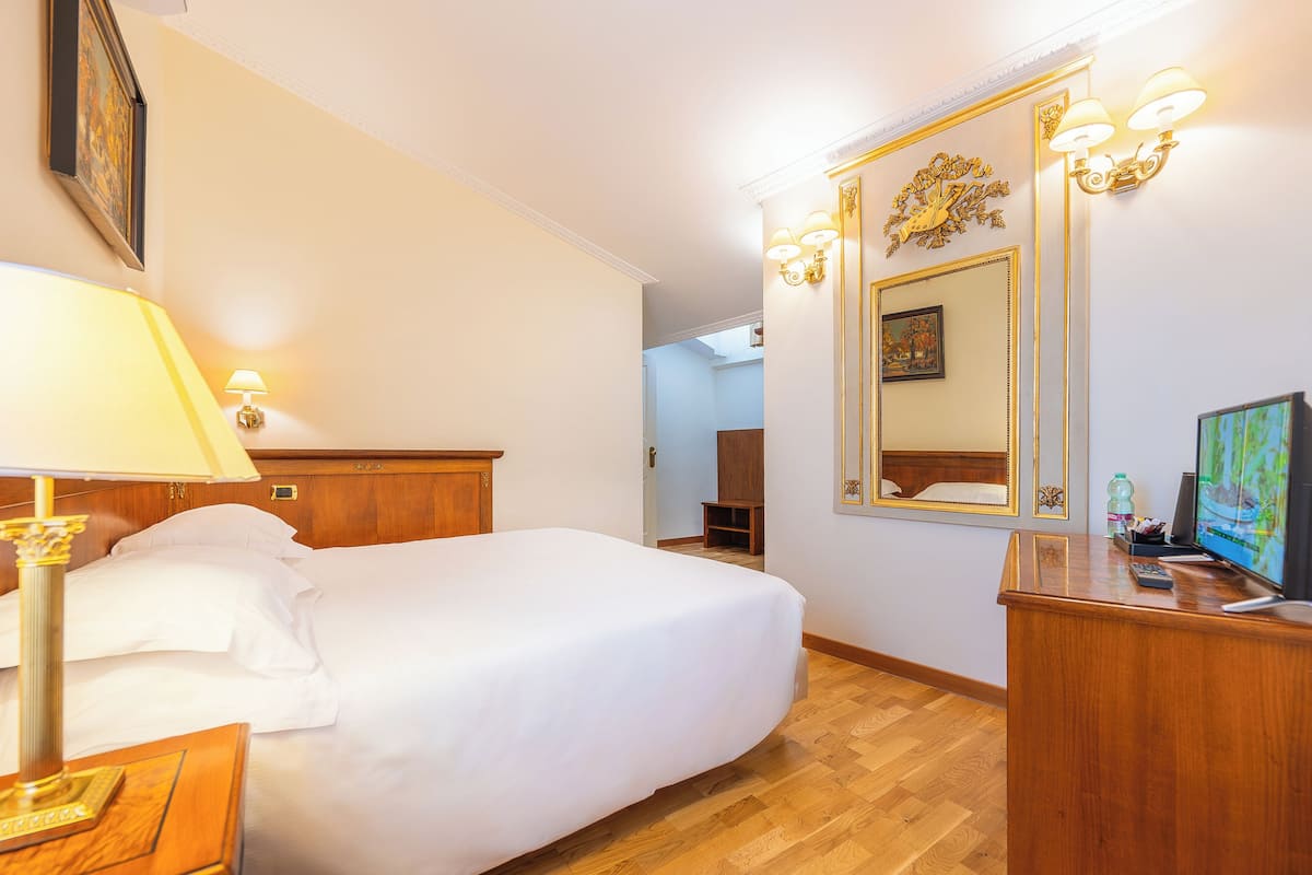 A hotel room with a bed, bedside lamp, wooden dresser, TV, and a mirror with wall sconces.