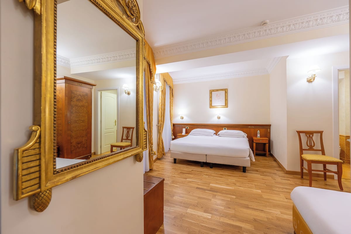 A hotel room with a large ornate mirror, a bed, a wooden wardrobe, a nightstand, and a chair.