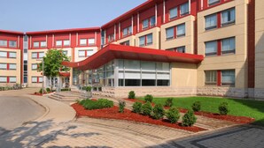 A multi-story building with a red roof and beige brick facade, featuring a central glass entrance and small landscaped areas with shrubs. - Residence & Conference Centre - Oakville (Oakville)