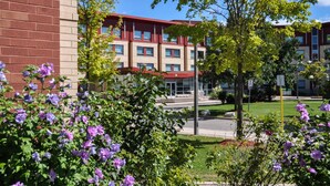 A multi-story building with a red roof and beige walls, surrounded by lush greenery and purple flowers. - Residence & Conference Centre - Oakville (Oakville)