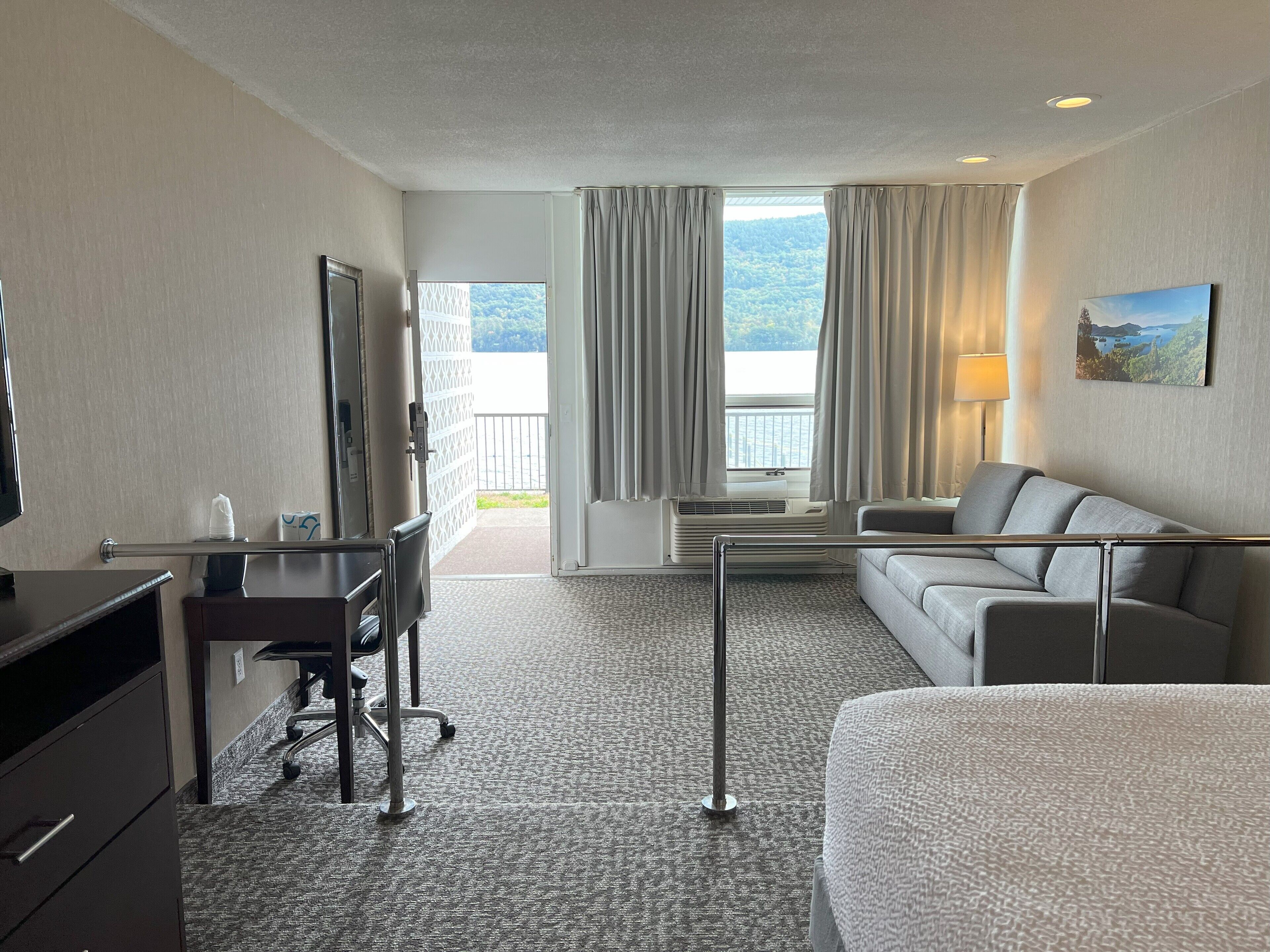 A hotel room with a bed, a desk with a chair, a sofa, a TV, and a window with curtains.