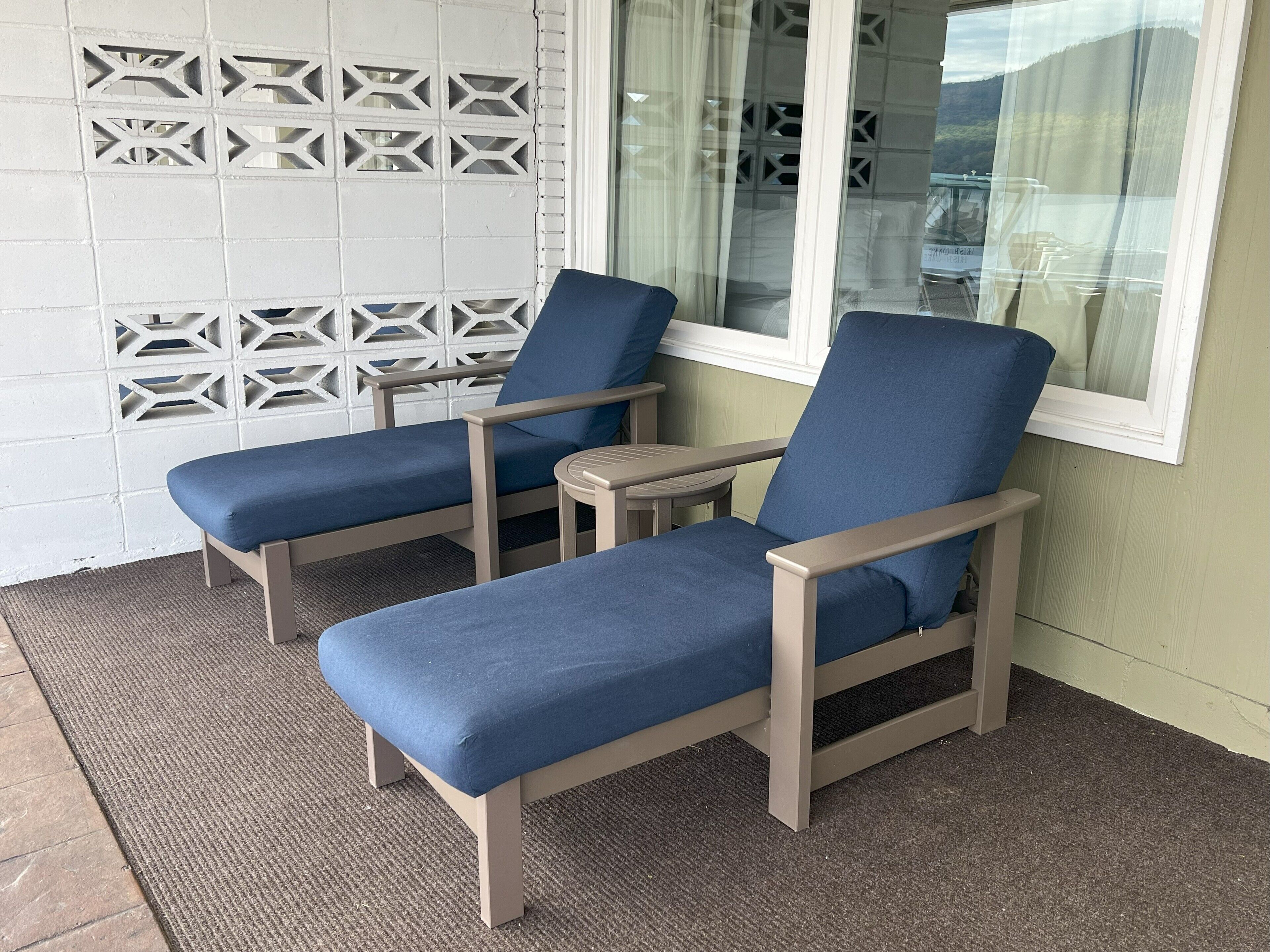Two blue cushioned chairs with wooden frames and a small table, positioned on a textured mat outside a building with a decorative white wall and a window with a view of the landscape.