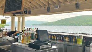 A bar area with a view of a lake and hills, a chalkboard menu, and a variety of bottles and glasses. - The Georgian Lakeside Resort (Lake George)
