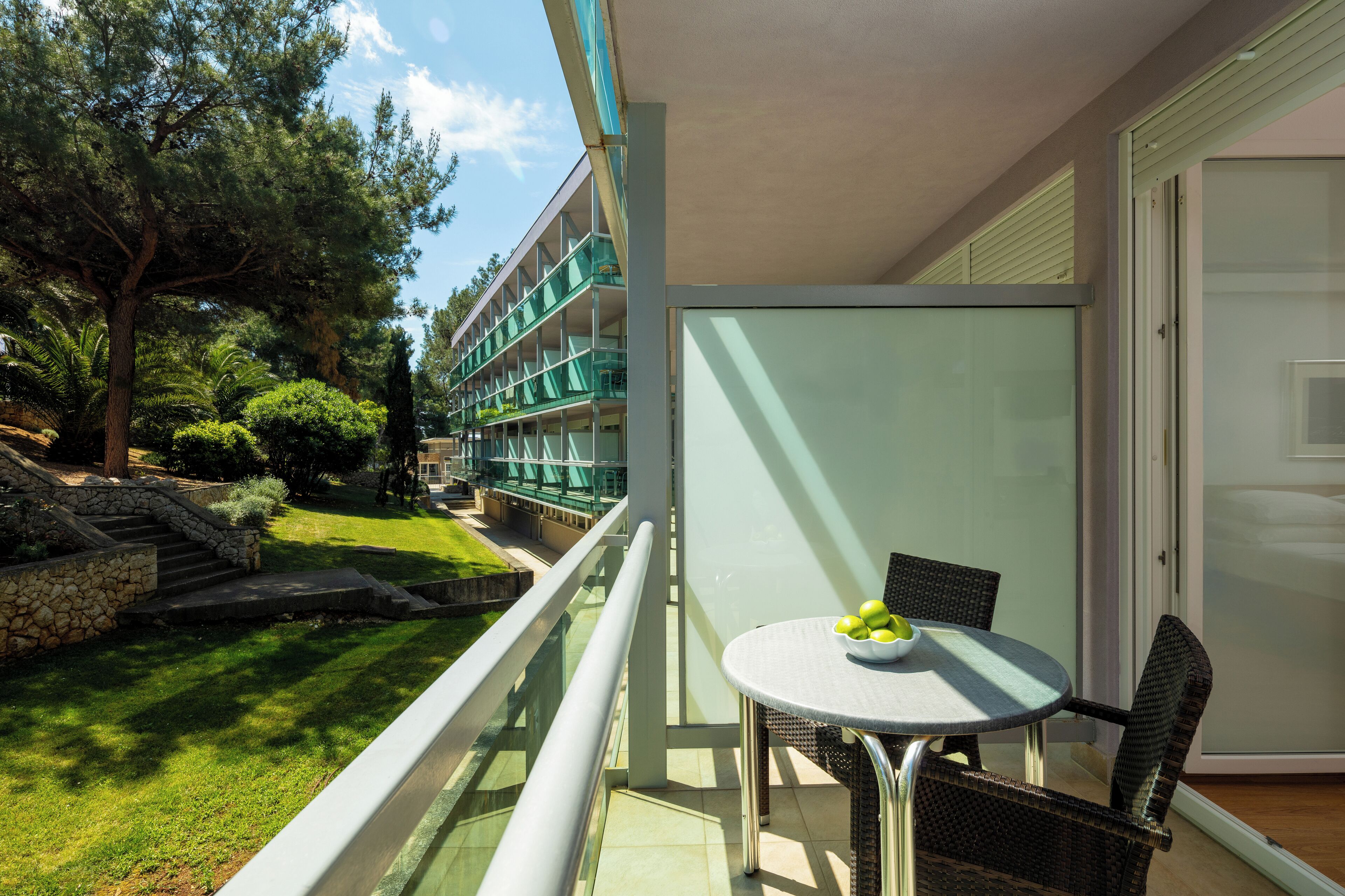 Un balcón con barandilla de vidrio, una mesa con sillas y vistas a un edificio con varios balcones.