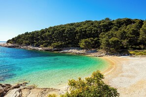 Uma praia costeira com águas turquesa cristalinas, areal arenoso e uma encosta arborizada ao fundo.