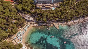 An aerial view of a coastal resort with a large building surrounded by dense trees, a sandy beach with umbrellas, and clear turquoise waters with boats. - Hotel Aurora (Mali Losinj)