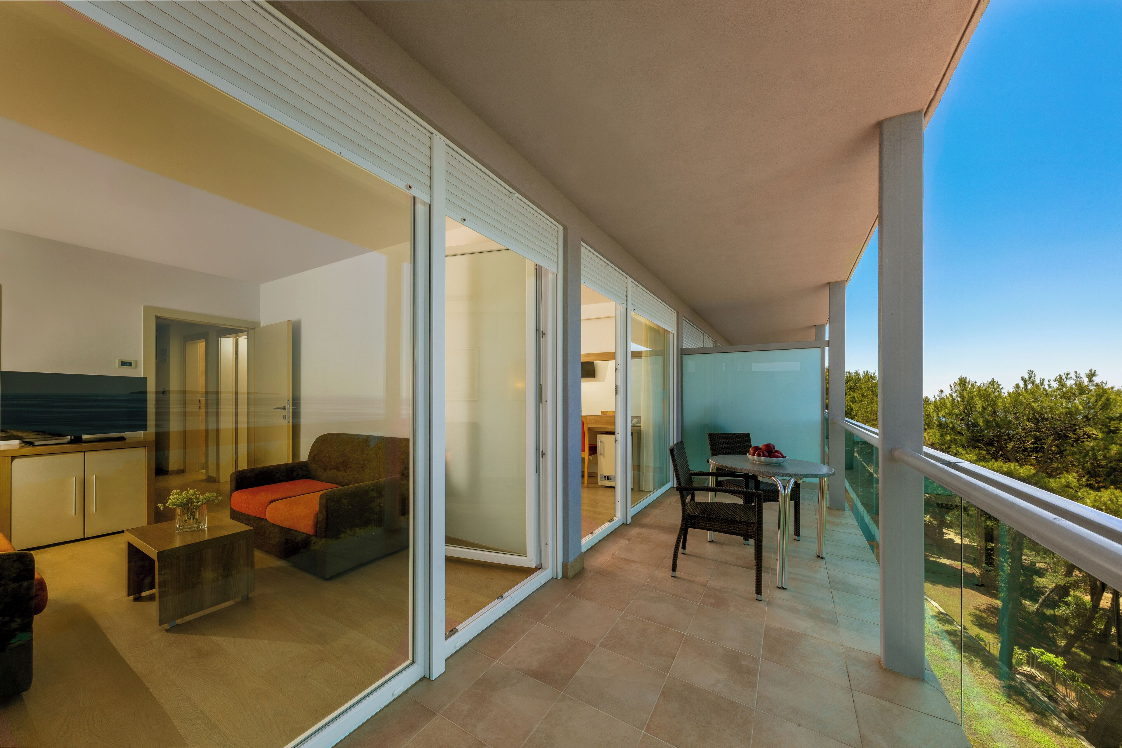 A modern living area with a sofa, coffee table, and a TV. There is a balcony with a table and chairs, and a glass door leading to another room.