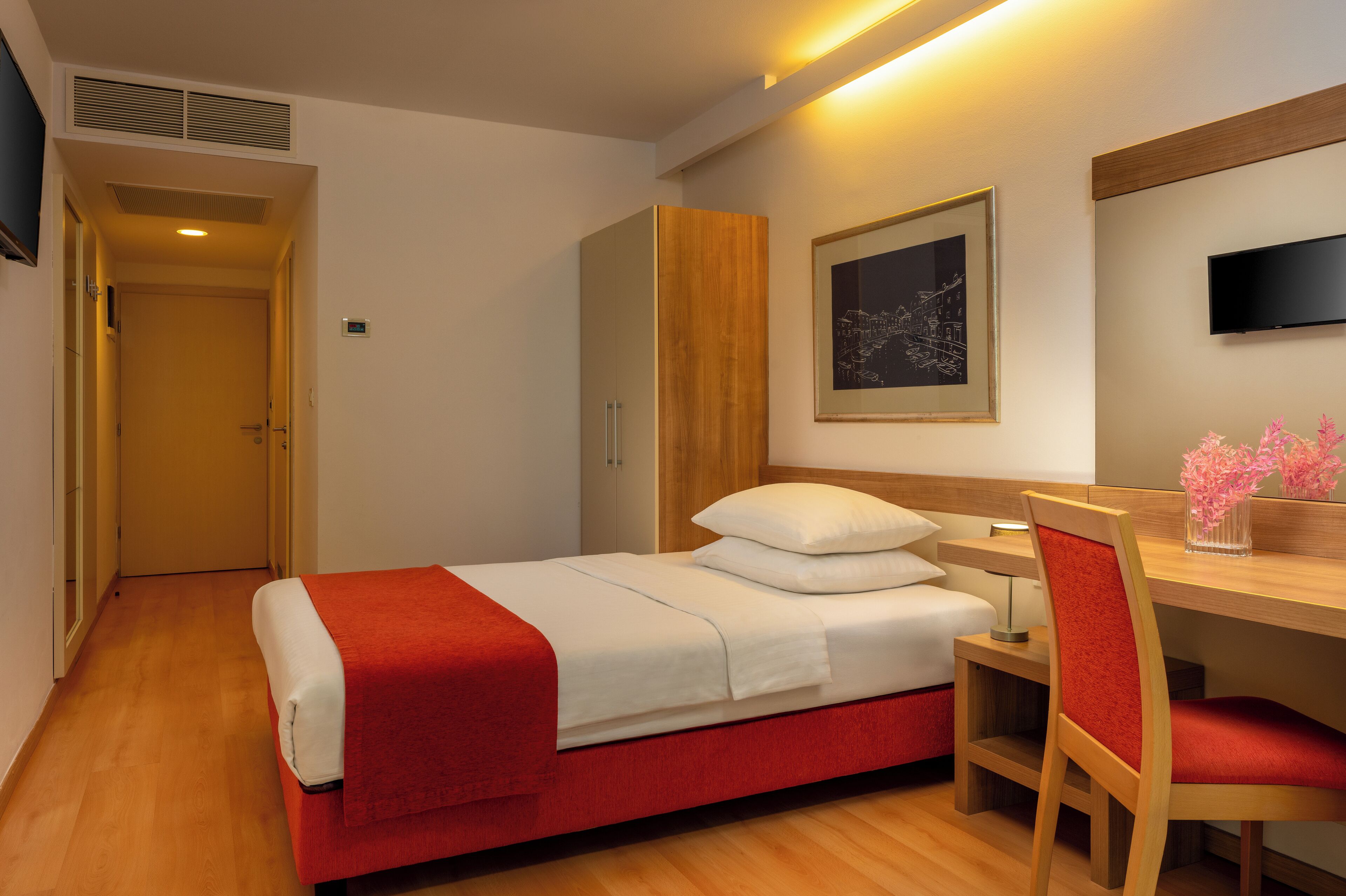 A hotel room with a bed, a desk, a chair, a wardrobe, a TV, and a framed picture on the wall.