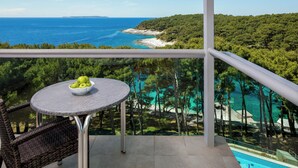 A balcony with a round table and wicker chairs, overlooking a turquoise sea and a forested hillside. - Hotel Aurora (Mali Losinj)