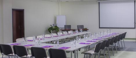 Un salón de conferencias con una larga mesa, sillas, botellas de agua y una pantalla de proyector.