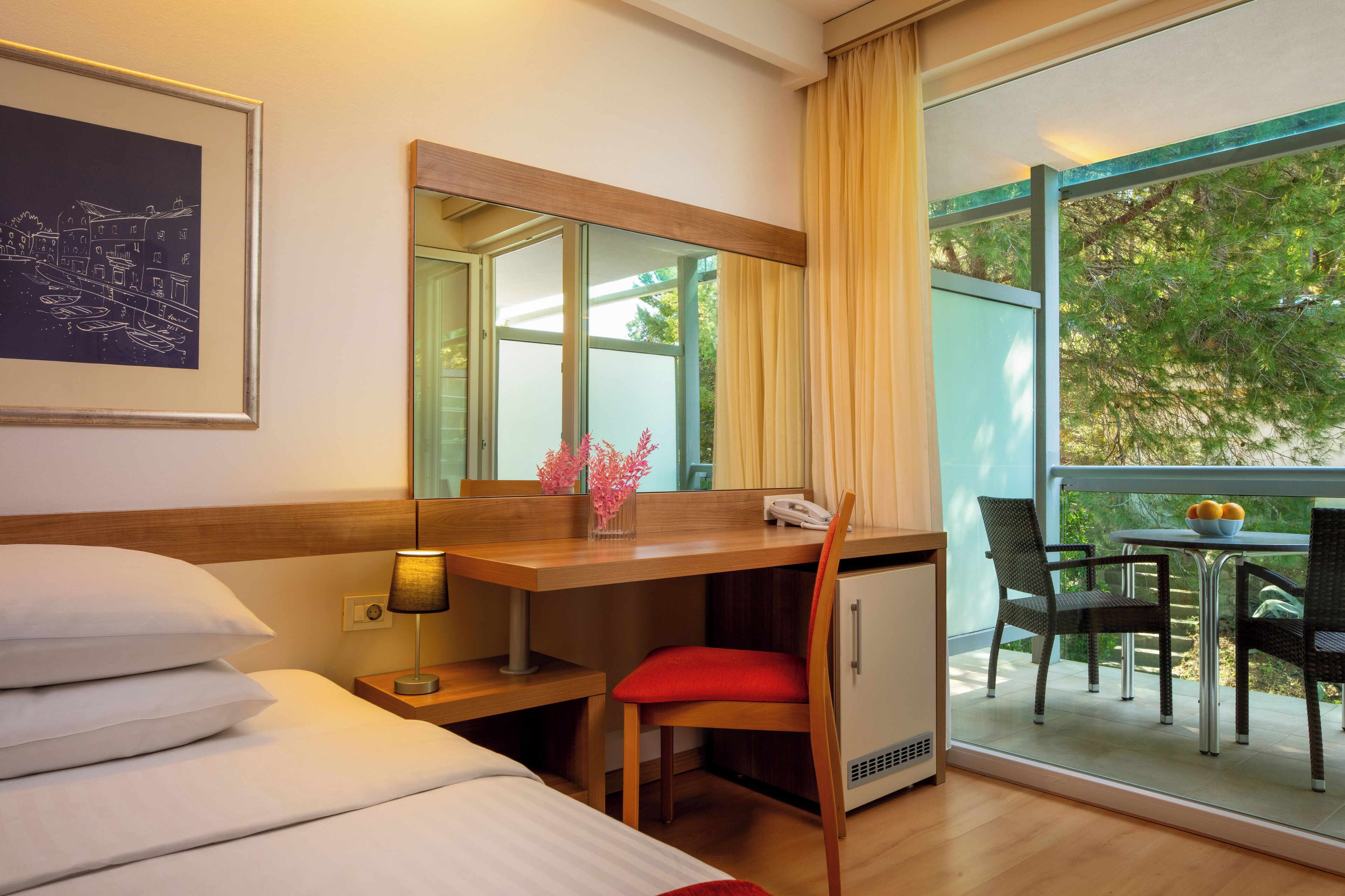 Une chambre d’hôtel comprenant un lit, un bureau avec une lampe, une chaise, un petit réfrigérateur, un miroir et un balcon avec une table et des chaises.