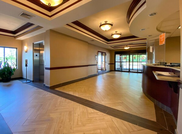 A spacious hotel lobby with a wooden herringbone floor, a reception desk, and a large glass door leading outside.