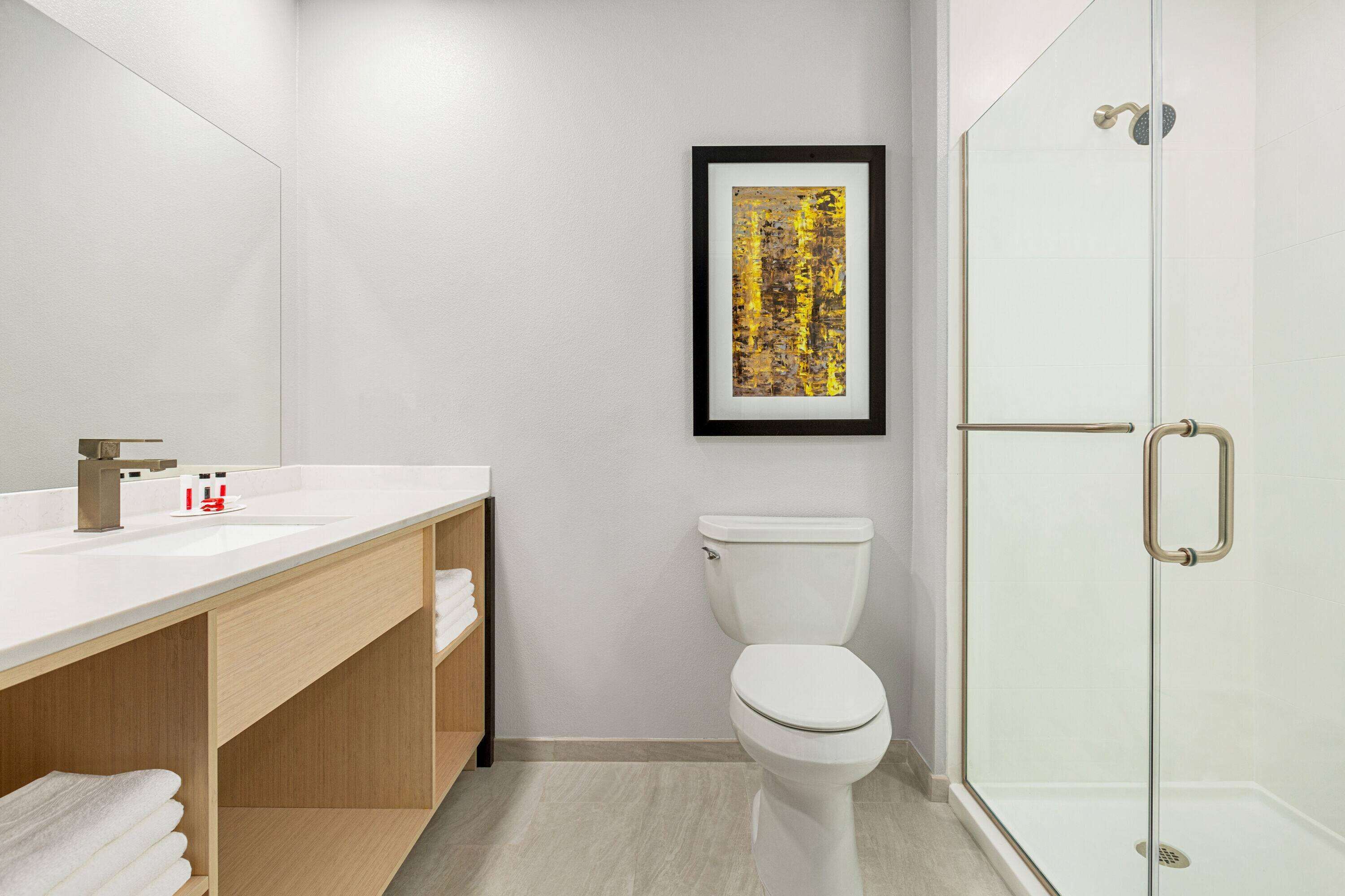 A modern bathroom with a white sink, wooden vanity, a framed abstract painting, a white toilet, and a glass shower enclosure.