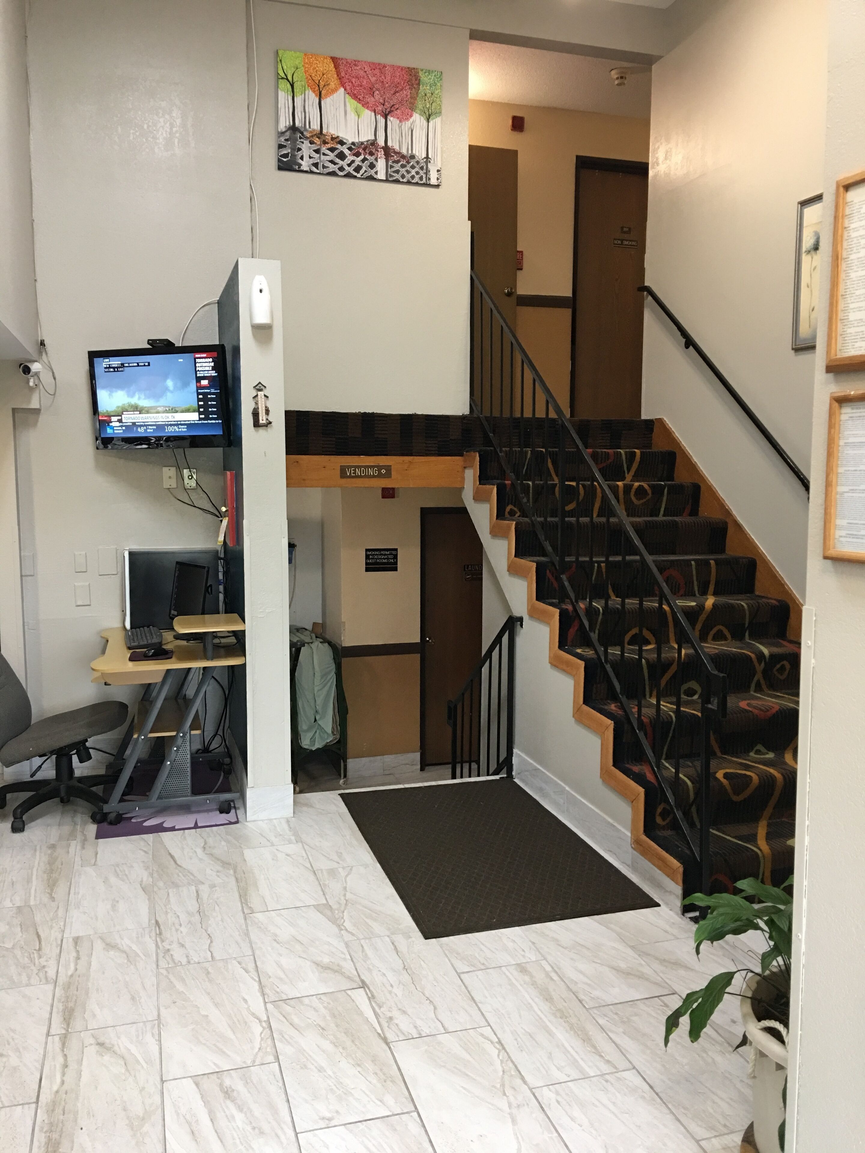 A hotel room with a desk, computer, and a TV mounted on the wall. There is a staircase with a sign indicating a 'Vending' area.