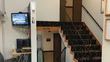 A hotel room with a desk, computer, and a TV mounted on the wall. There is a staircase with a sign indicating a 'Vending' area.