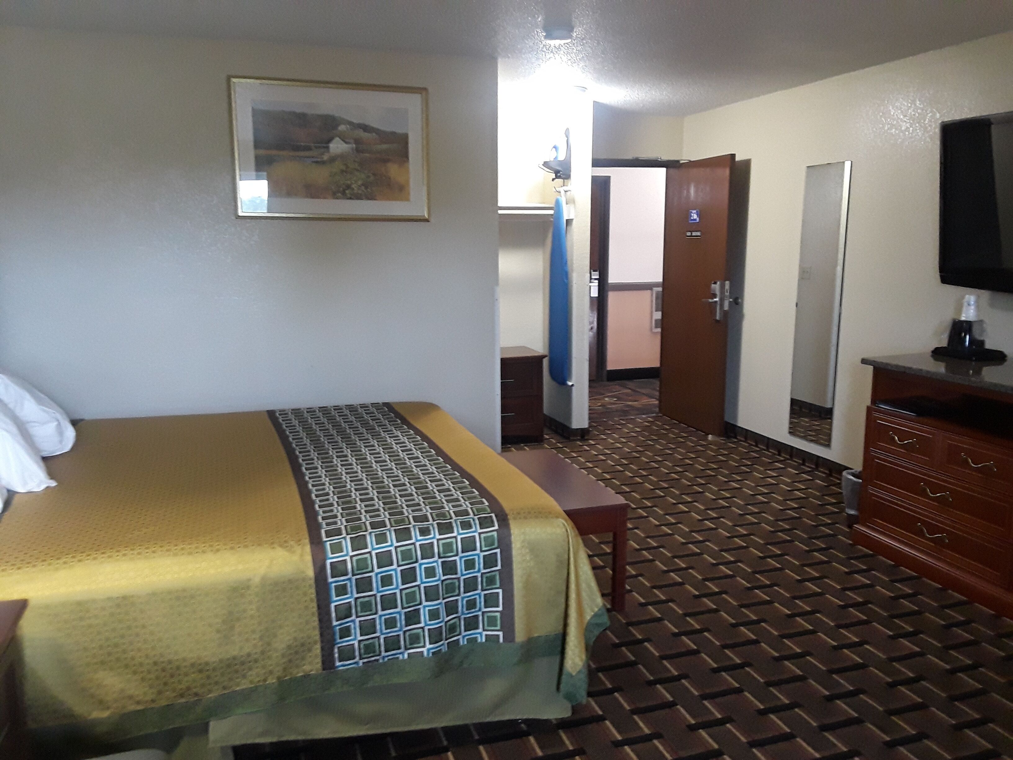 A hotel room with a bed, a dresser, a mirror, a television, and a door leading to another room.