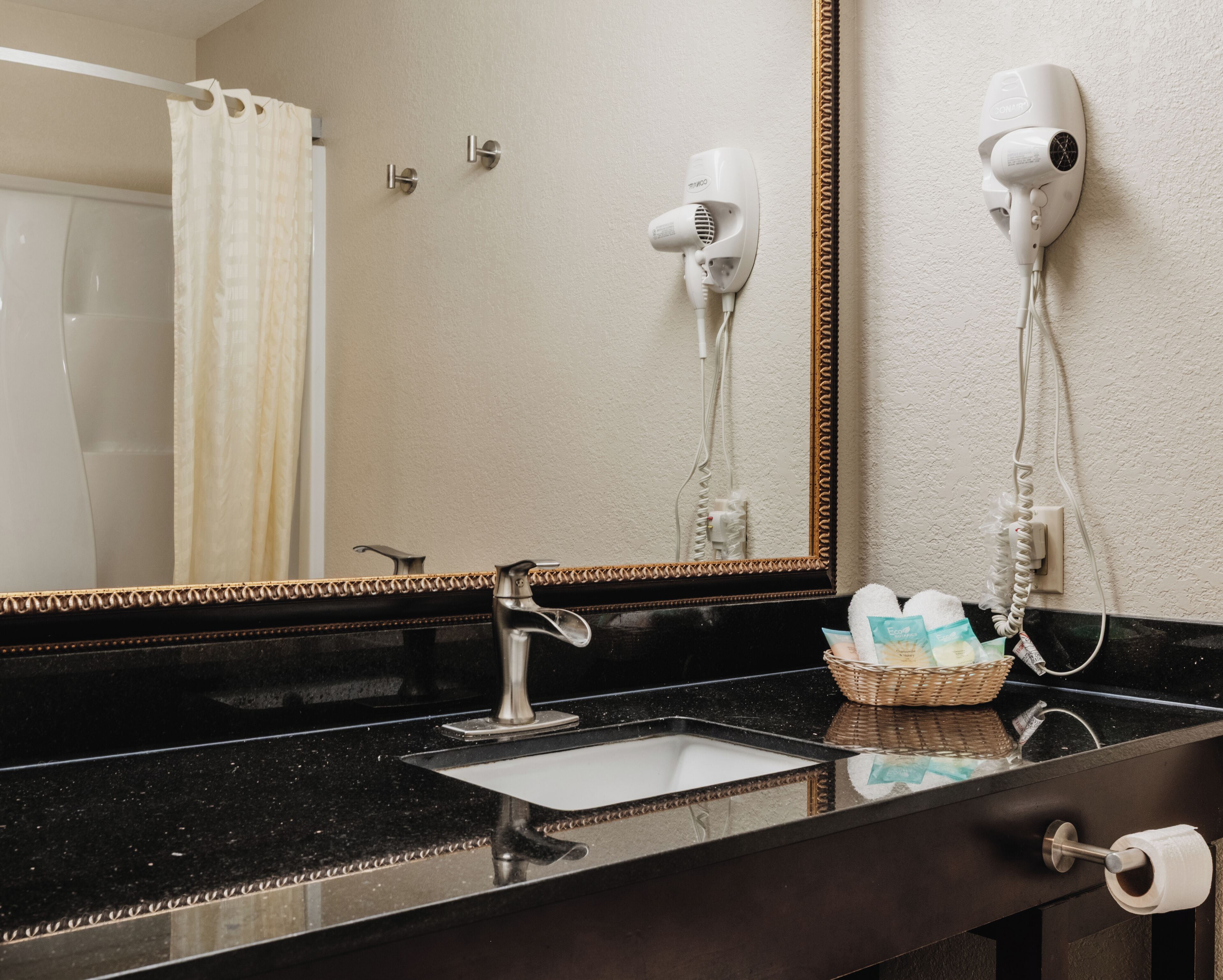 A bathroom with a black countertop, a sink, a mirror with a gold frame, and two hair dryers mounted on the wall.