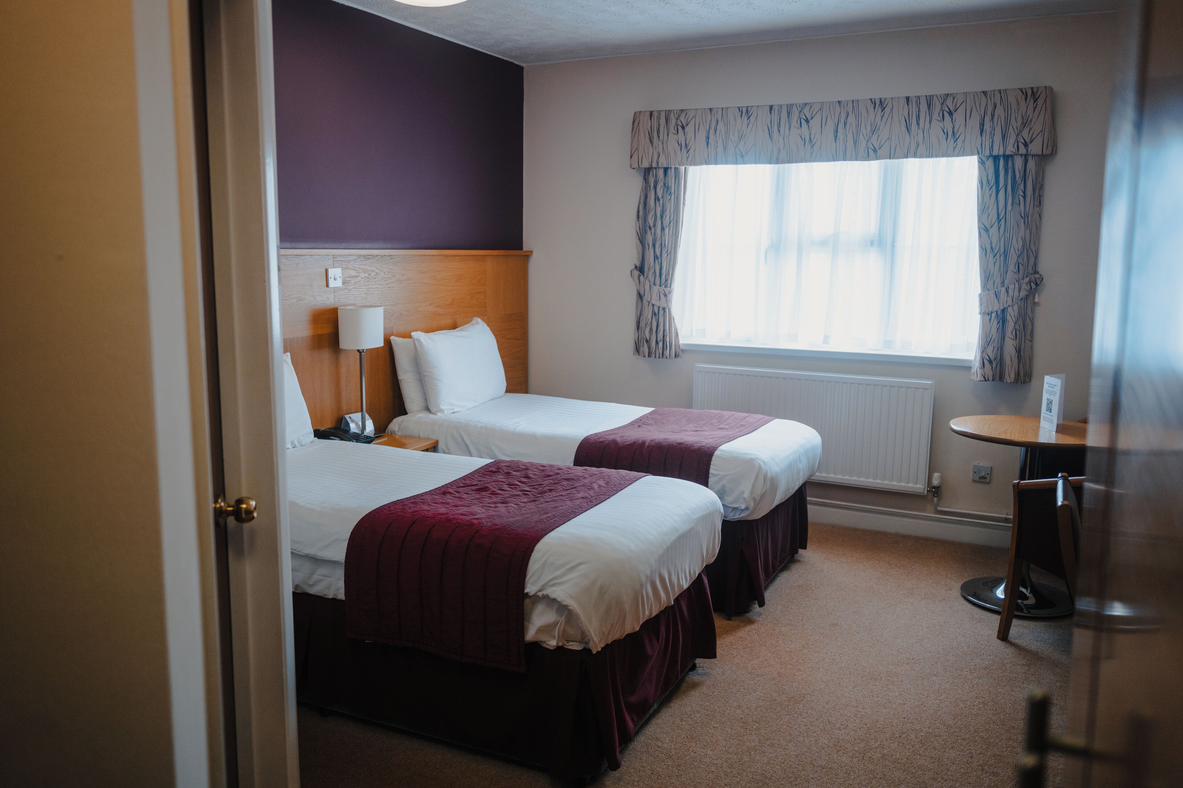 A hotel room with two beds, a wooden headboard, a window with curtains, a radiator, and a small table with a chair.