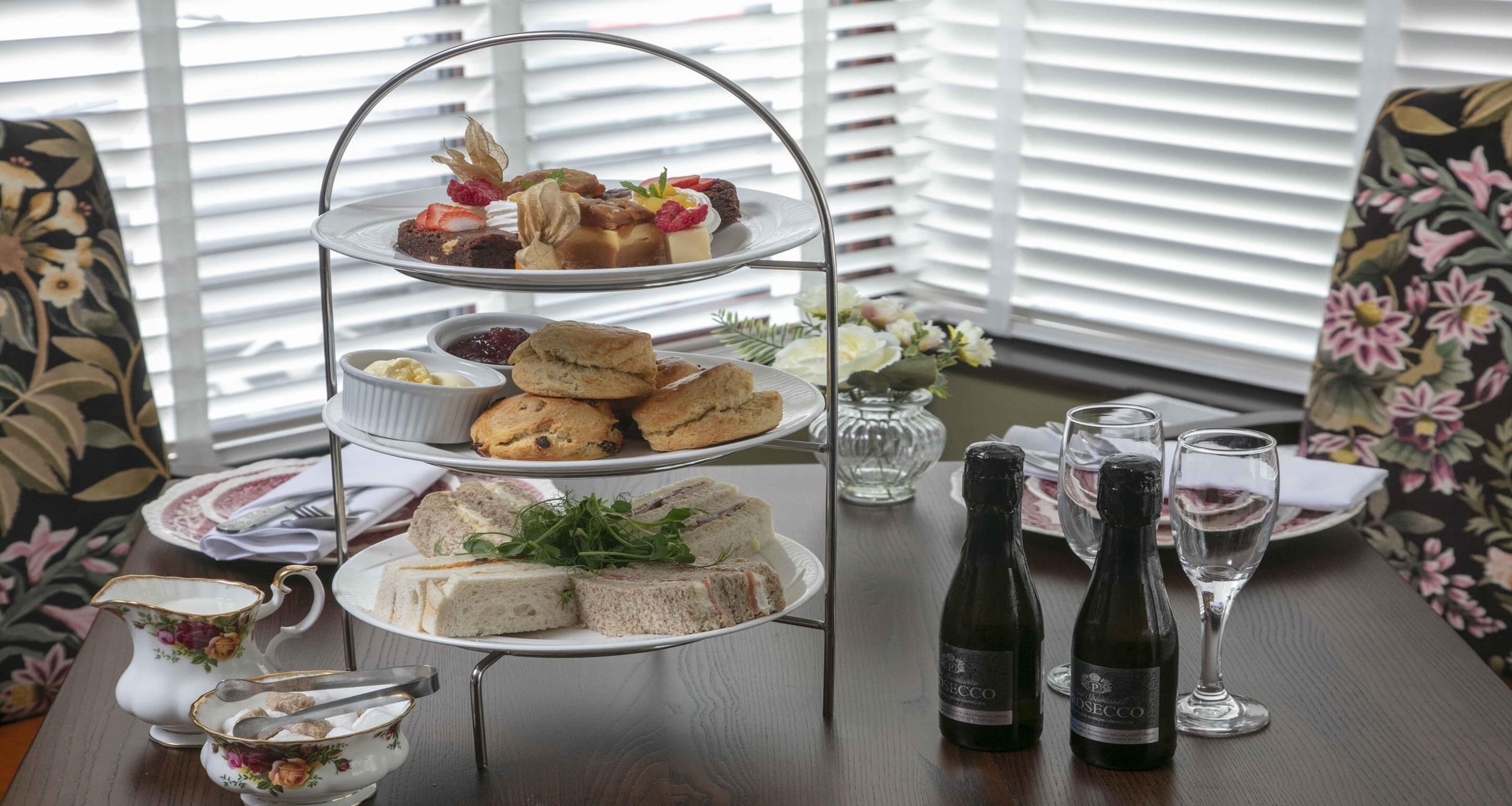 Un soporte de tres niveles con diversos pasteles y sándwiches, dos botellas de vino y una silla con estampado floral.