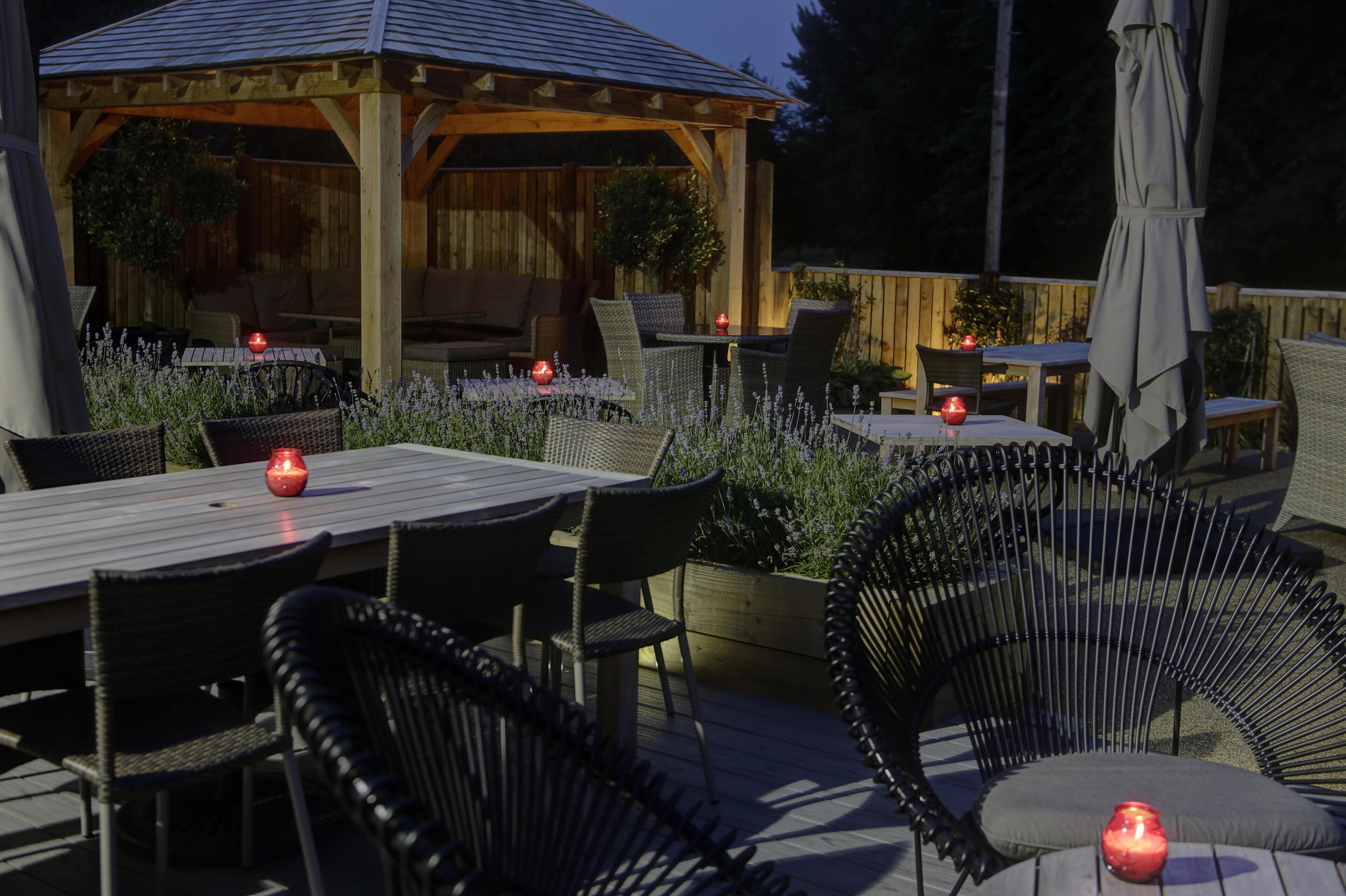 An outdoor patio area with wooden tables and chairs, surrounded by plants and a wooden pergola.