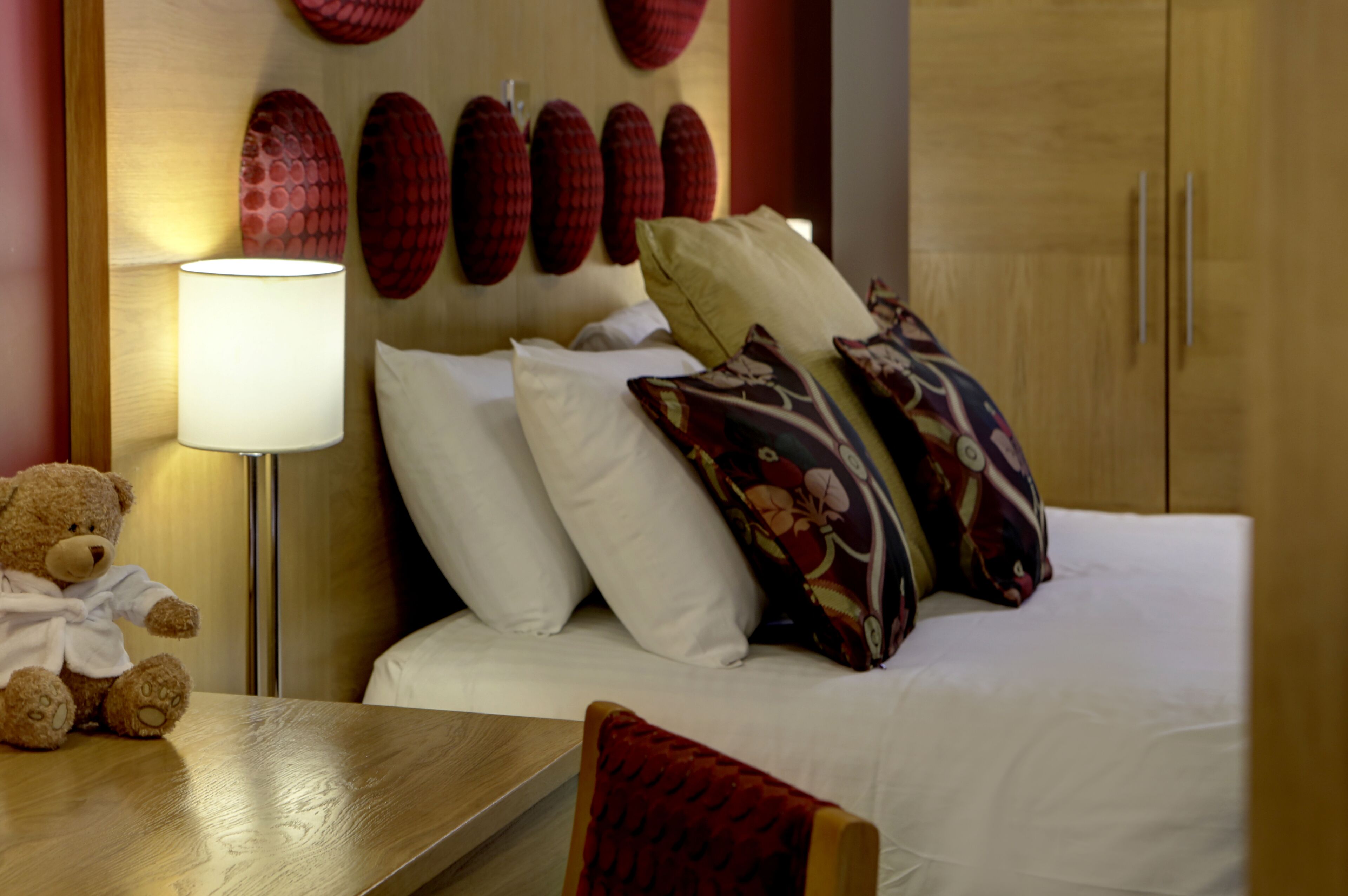 Ein ordentlich bezogenes Bett mit weißen und gemusterten Kissen, einer Nachttischlampe und einem Teddybären auf einem Holznachttisch.