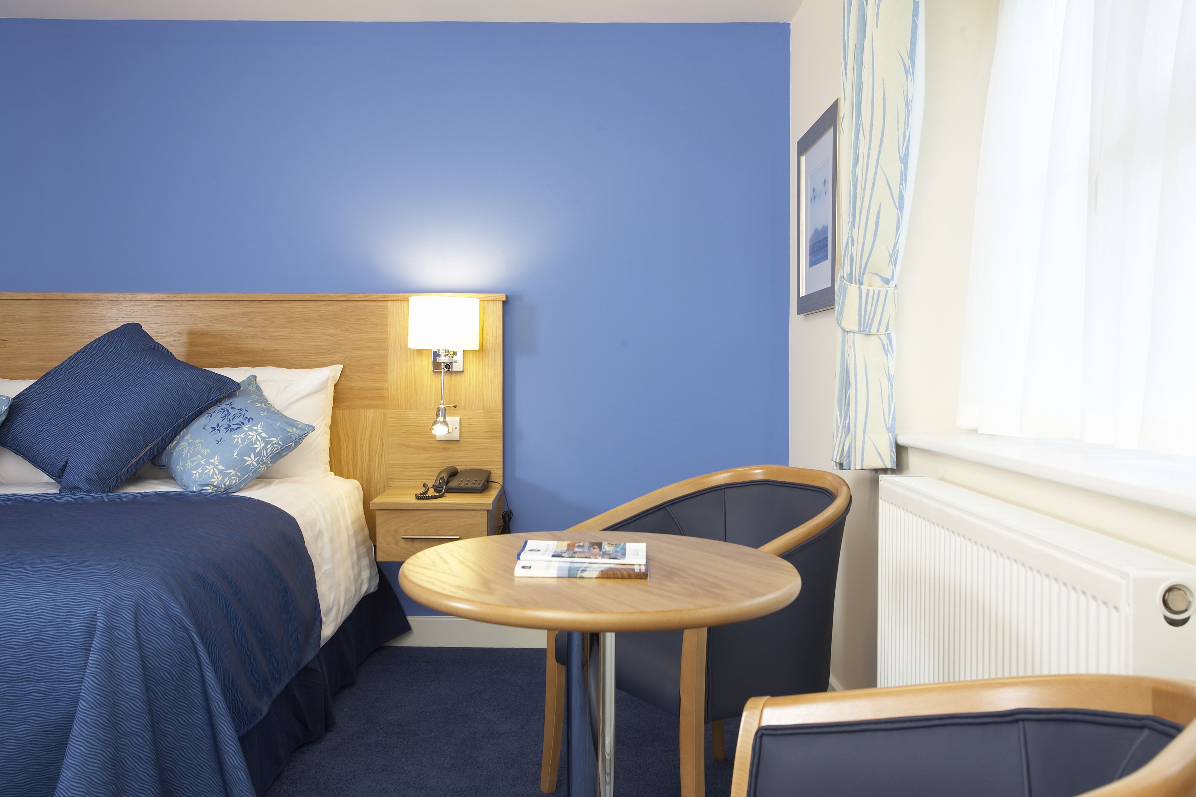 Ein Hotelzimmer mit Bett, Nachttisch, Lampe, Stuhl und einem kleinen Tisch mit einer Zeitschrift.