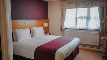 Hotelkamer met een groot bed, een houten hoofdbord, een nachtkastje met lamp, een raam met gordijnen en een radiator.
