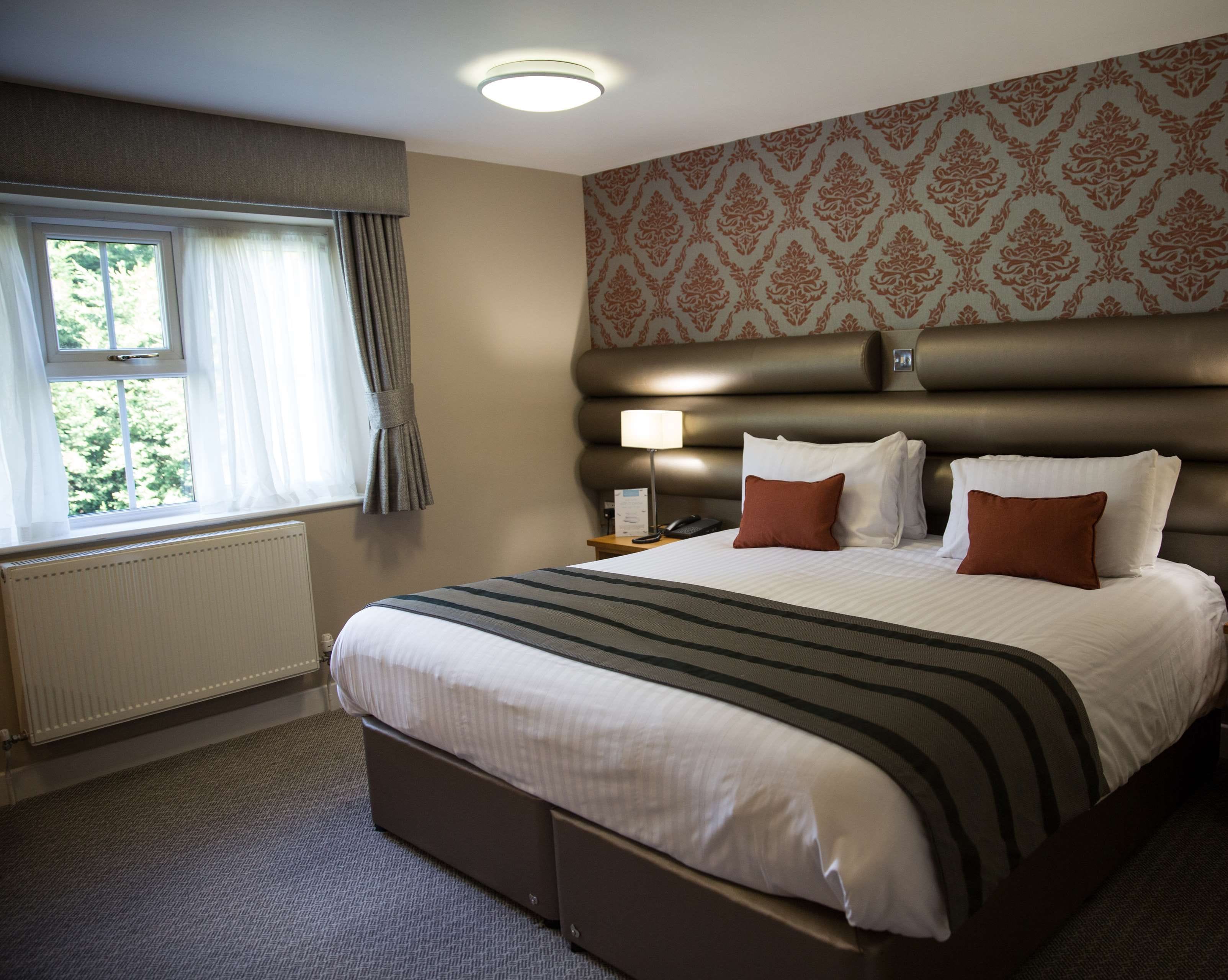 A hotel room with a large bed, a bedside table with a lamp, a window with curtains, and a patterned wallpaper.