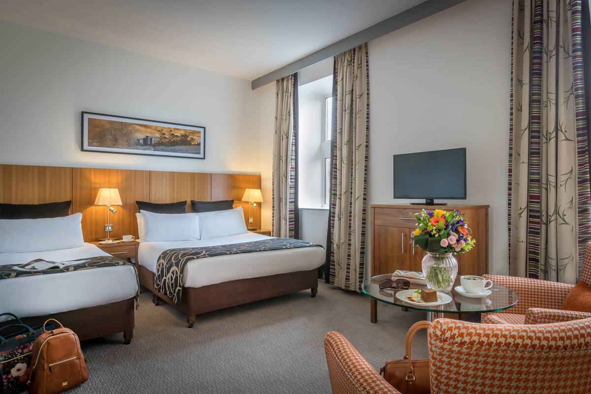 A hotel room with two beds, a TV, a coffee table with a vase of flowers, and a view of the outdoors.