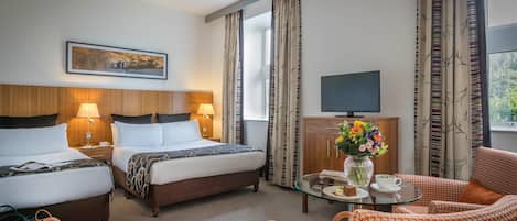 Habitación de hotel con dos camas, televisión, una mesa de centro con un florero, y vista al exterior.