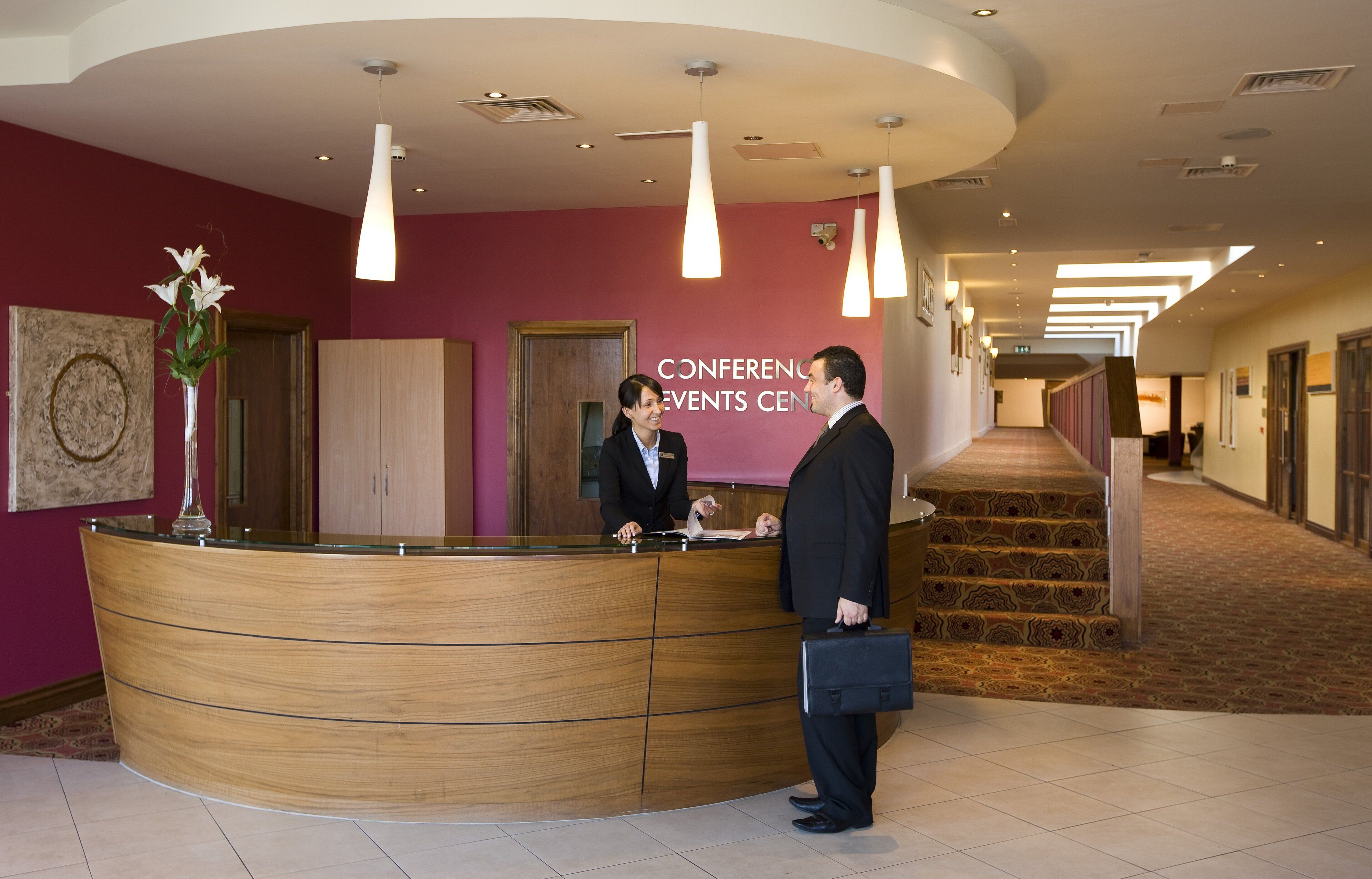 Zona de recepción con un mostrador curvo de madera, una recepcionista y un hombre con maletín.
