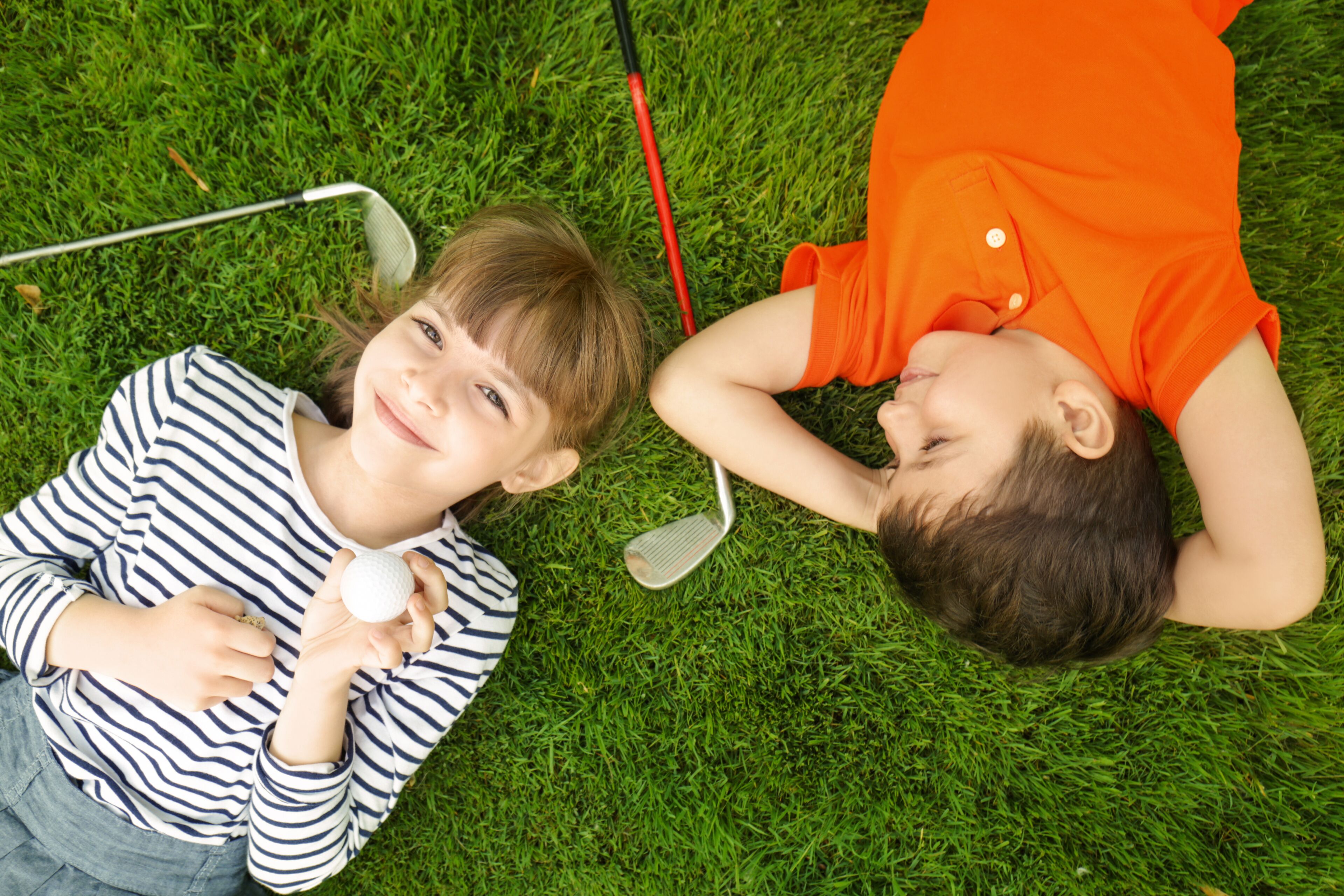 Two children lying on grass, one holding a golf ball and the other holding a golf club.