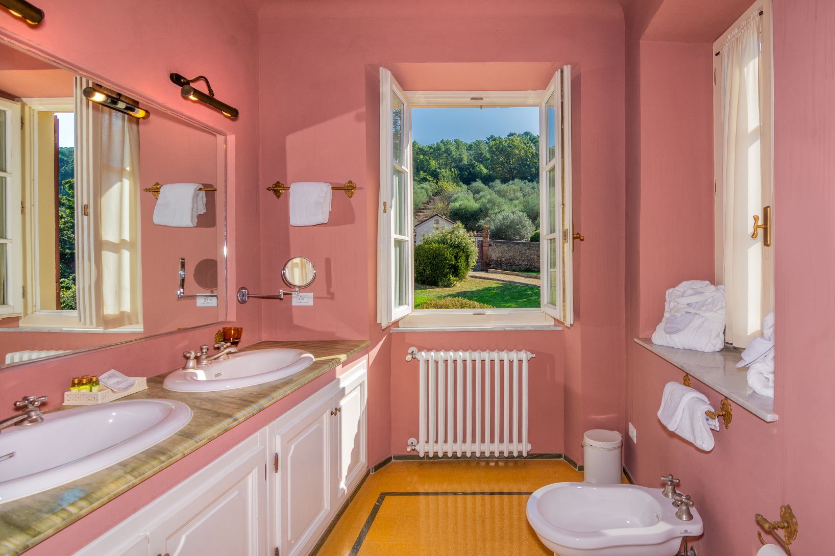 Un bagno rosa con due lavandini, un water e una finestra che si affaccia sul verde.