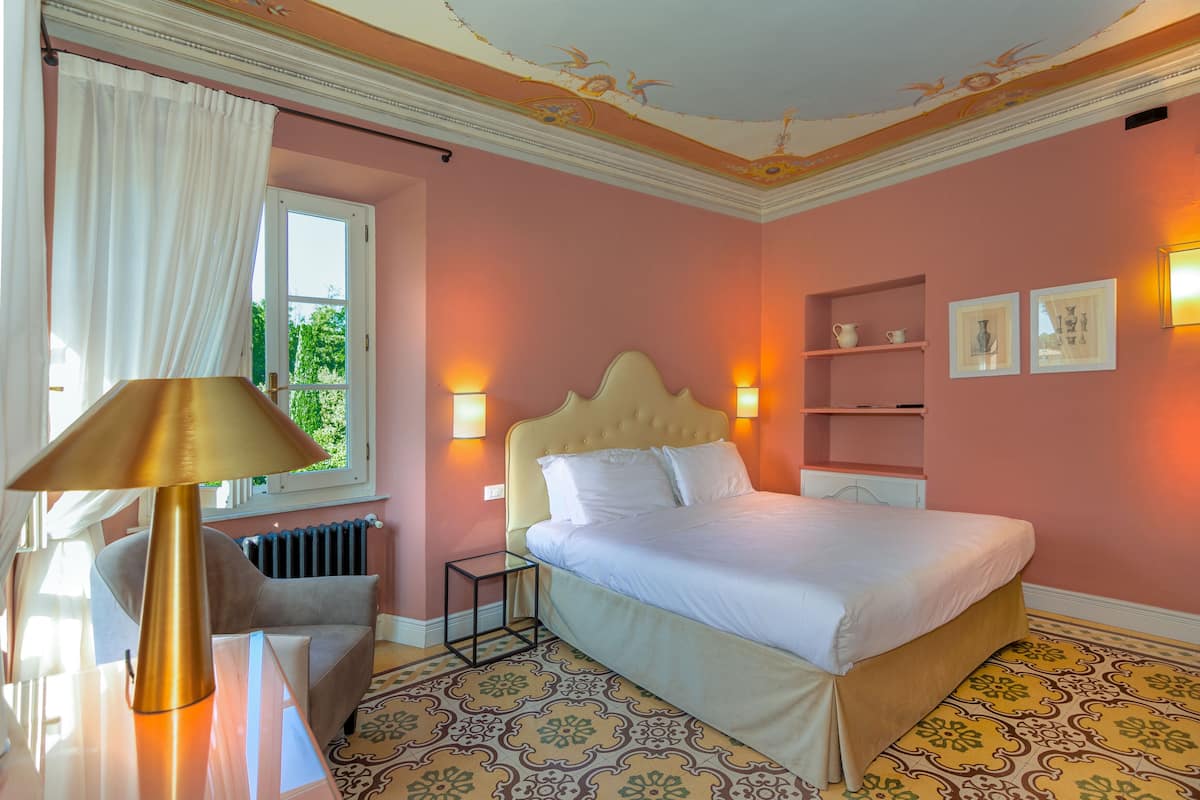 A bedroom with a bed, bedside lamps, a nightstand, a chair, a window with curtains, and a radiator.