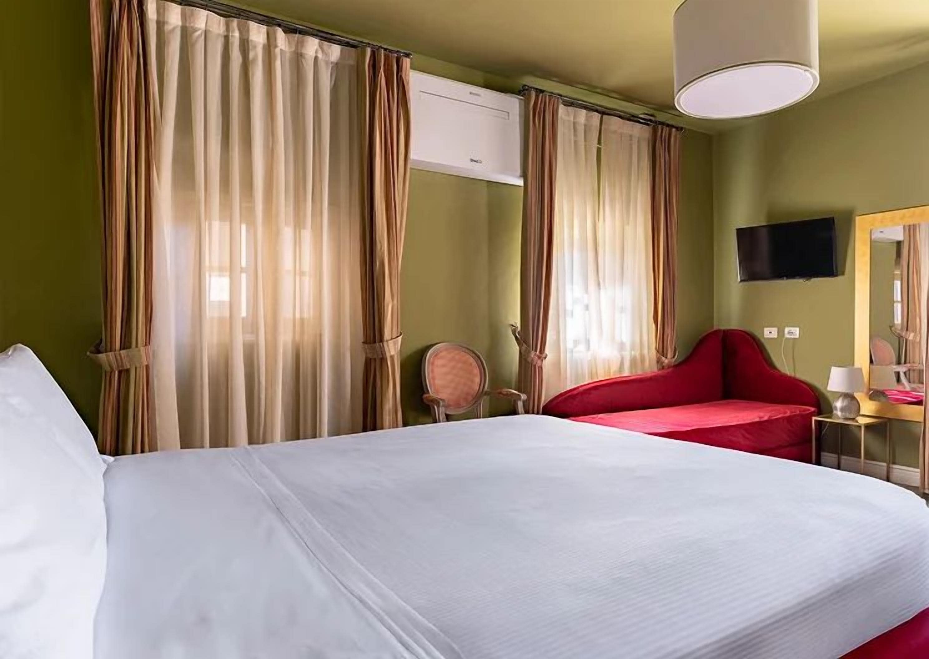 Une chambre d’hôtel avec un grand lit, un fauteuil rouge, un bureau avec une lampe et des rideaux.