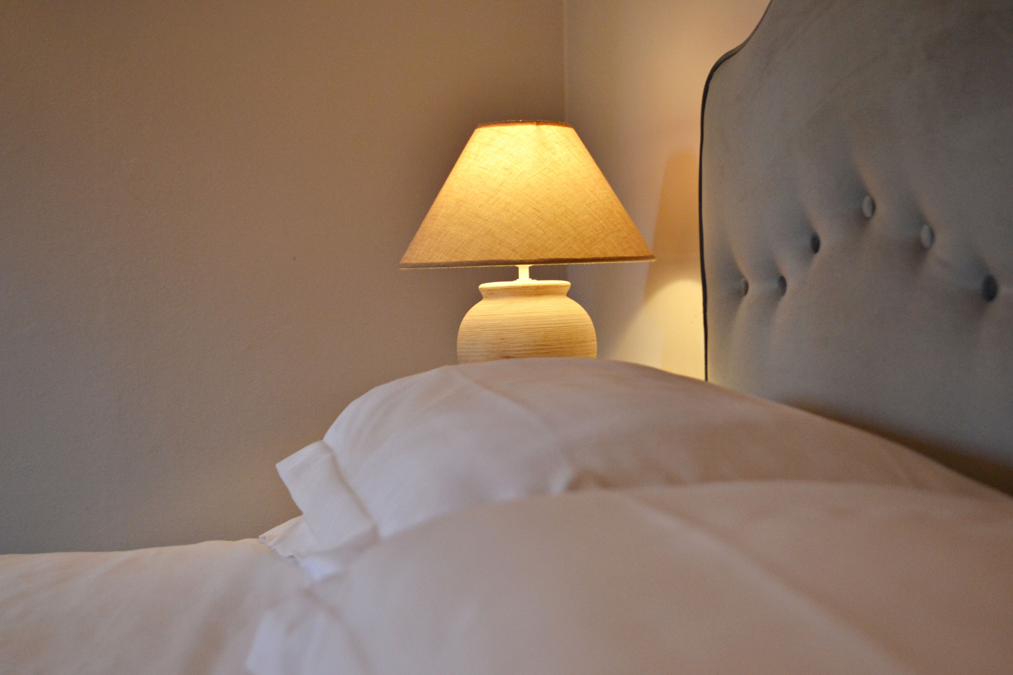 Een net opgemaakt bed met witte lakens en een lamp met een beige lampscherm op een nachtkastje.