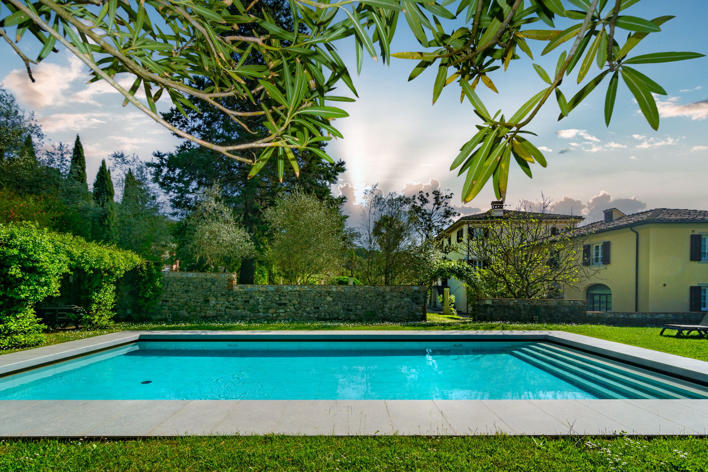 A rectangular swimming pool with a stone border, surrounded by a well-maintained lawn and a stone wall.