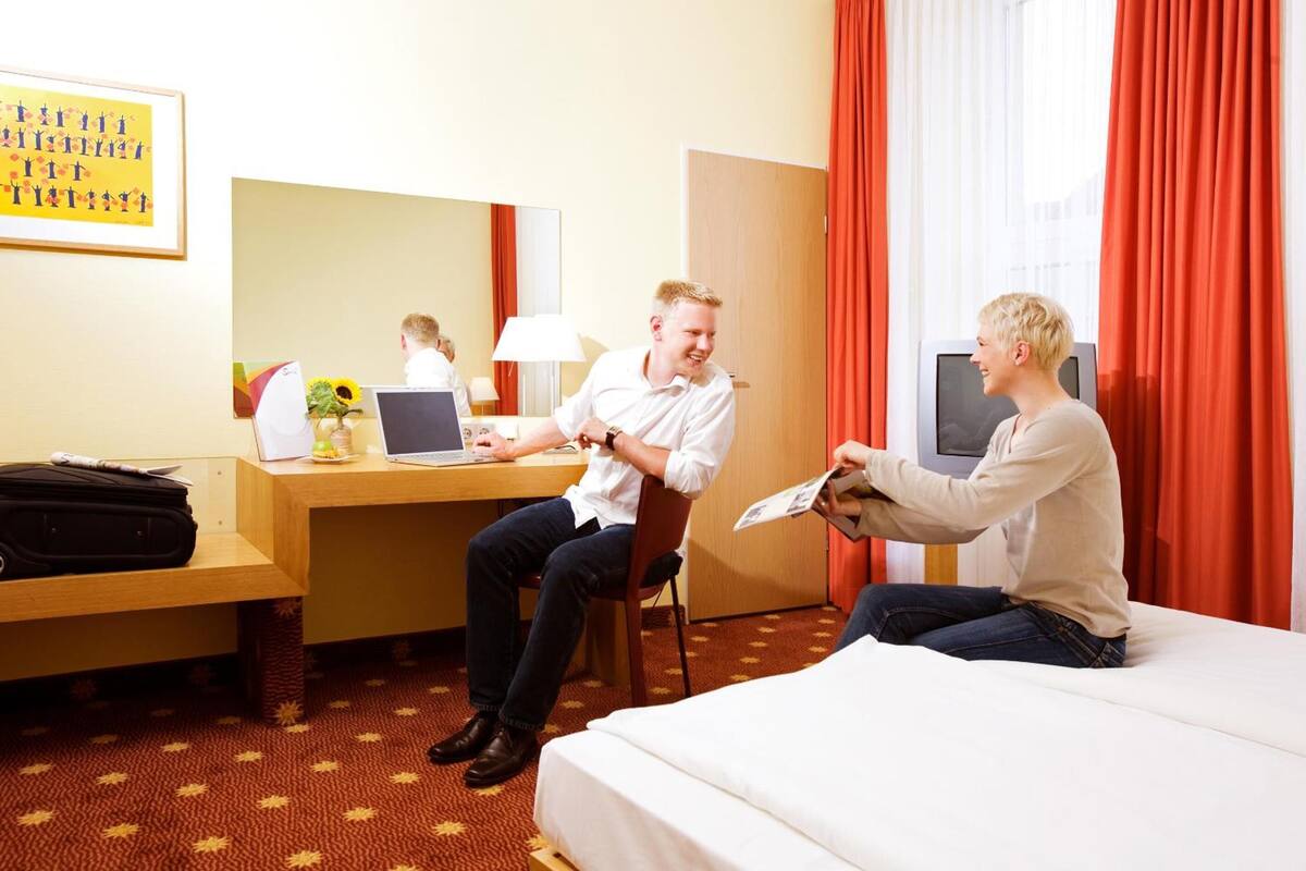 Ein Hotelzimmer mit einem Bett, einem Schreibtisch, einem Laptop, einem Sessel und einem Fernseher.