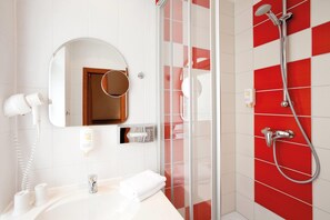 A bathroom with a white sink, a mirror, a hairdryer, a shower with a red and white tiled wall, and a glass shower door. - Vienna House Easy by Wyndham Rostock (Rostock)