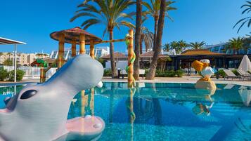 Ein Swimmingpool mit einer Delfin-Skulptur, Palmen und einem Gebäude mit einem Reetdach im Hintergrund.