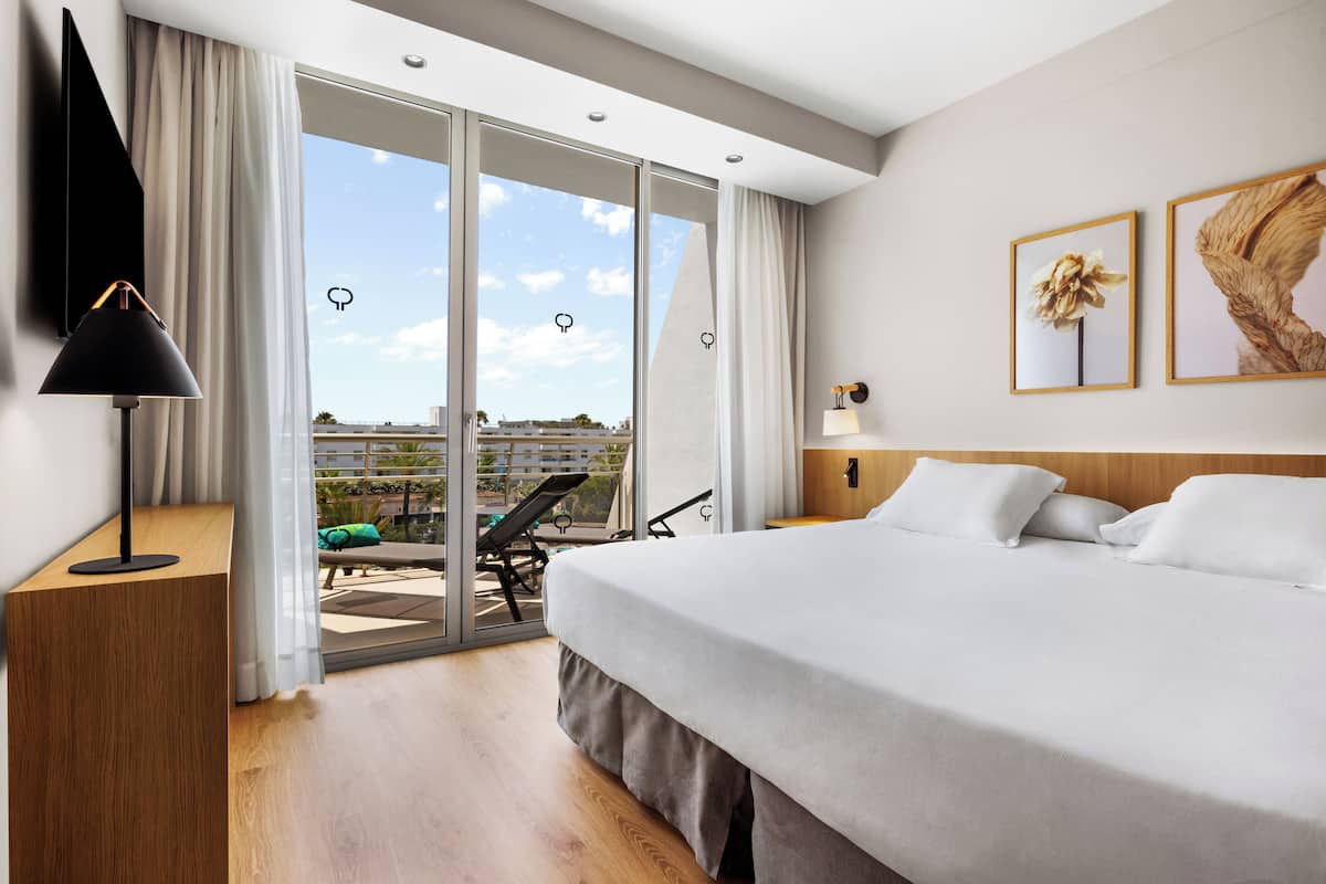 A hotel room with a large bed, a TV, a desk, and a balcony with a view of the city.