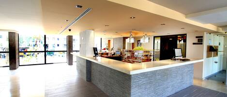 A spacious hotel lobby with a central reception desk, modern lighting fixtures, and a view of the outdoor area through large windows.
