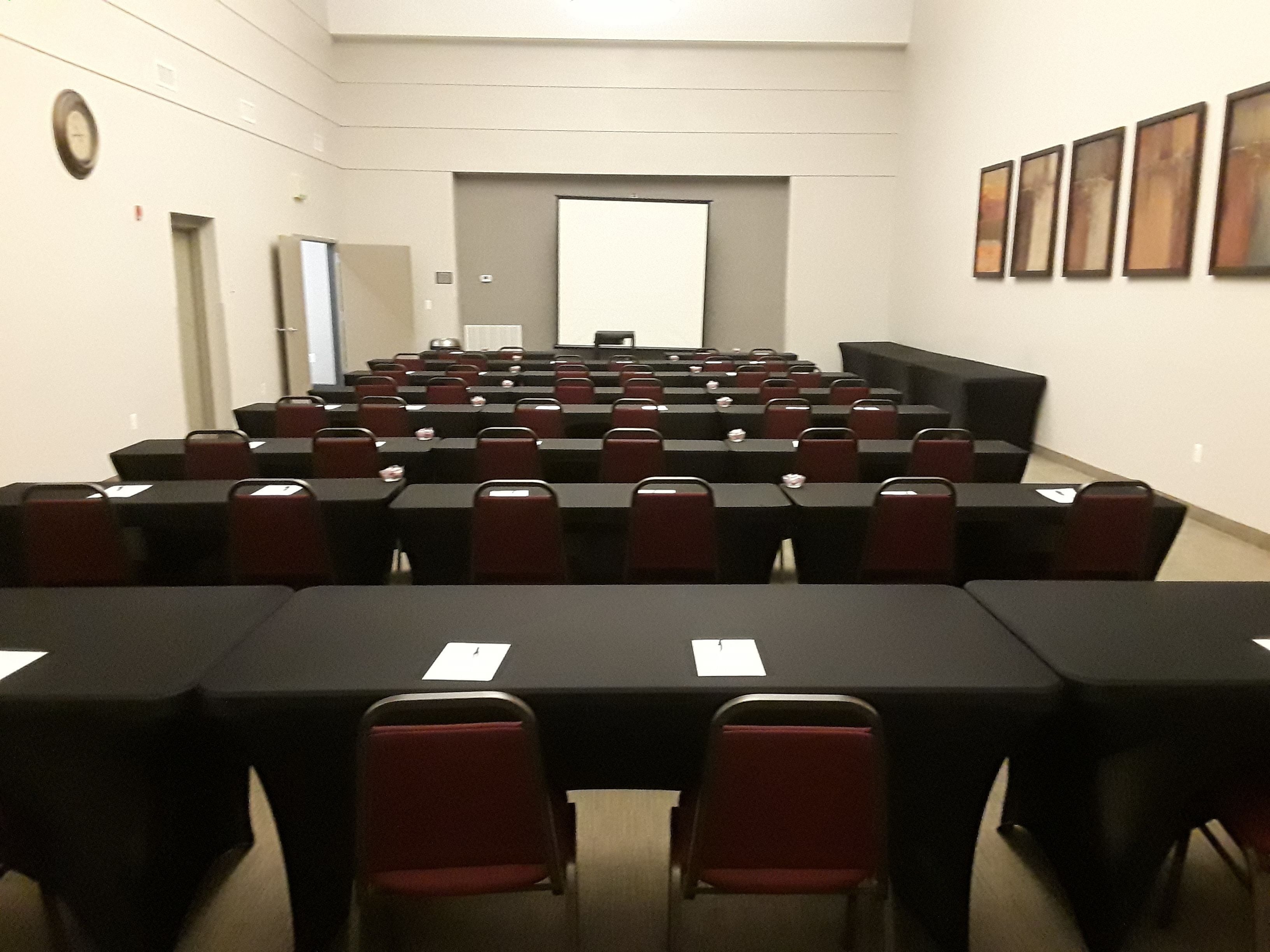 A conference room with rows of tables and chairs, a projector screen, and framed artwork on the walls.