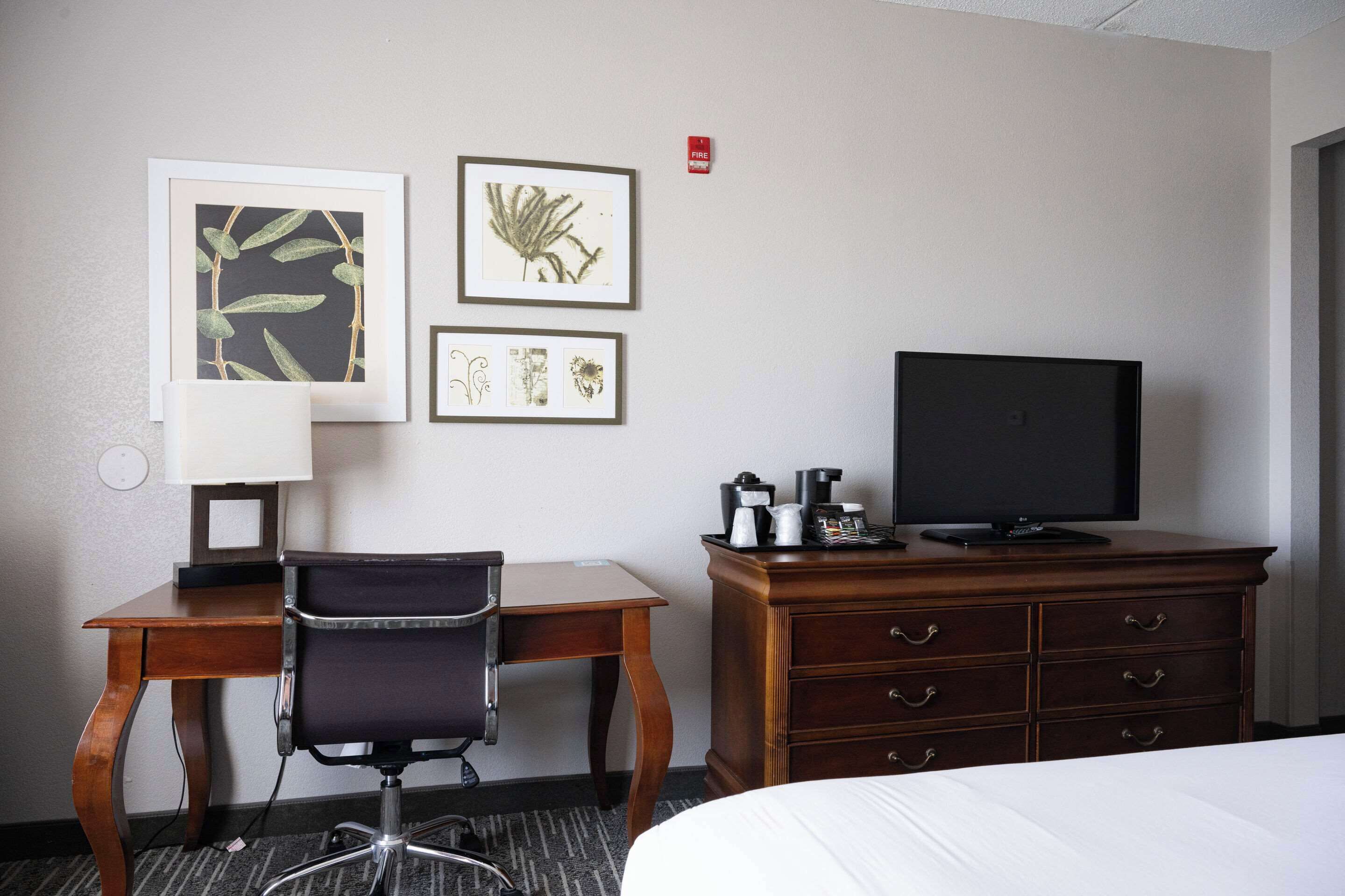 Une chambre d’hôtel comprenant un lit, un bureau avec une chaise, une télévision, une commode et des œuvres d’art encadrées accrochées au mur.