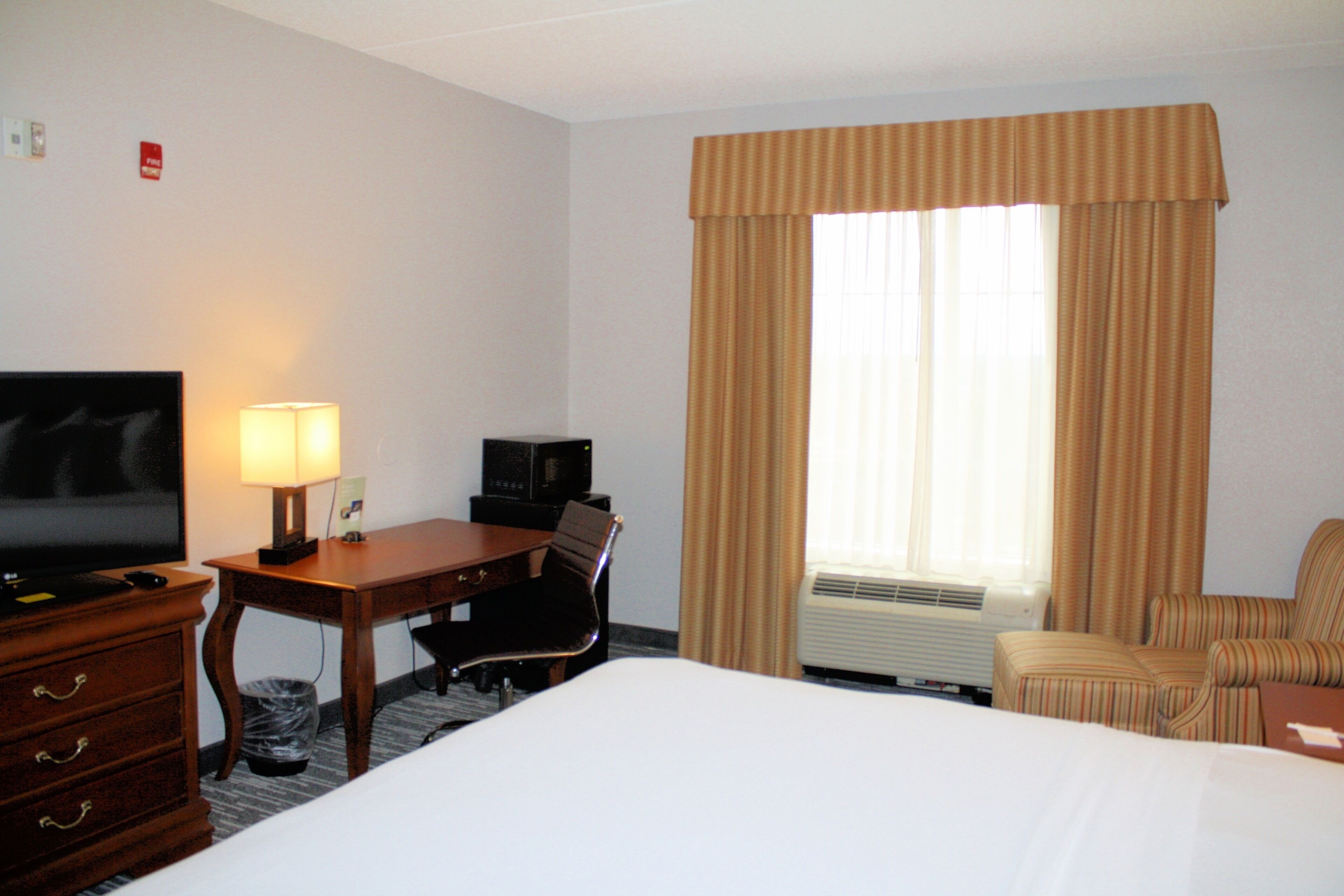 Een hotelkamer met een bed, een bureau met lamp, een televisie, een stoel en een raam met gordijnen.